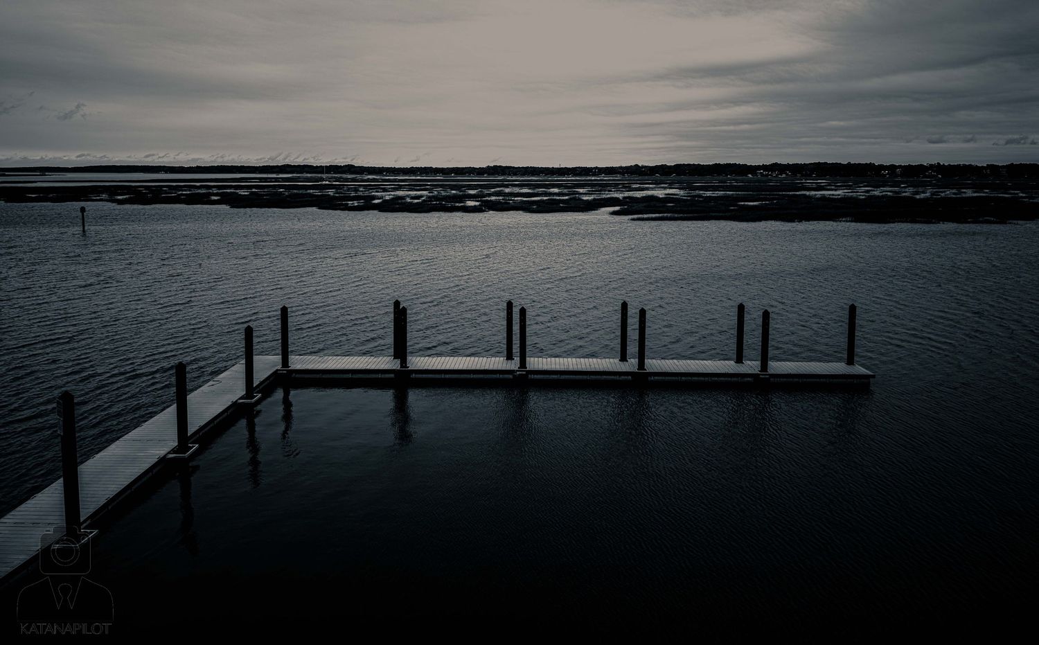 Moody black and white seascape with wooden dock posts silhouetted against a dramatic cloudy sky.