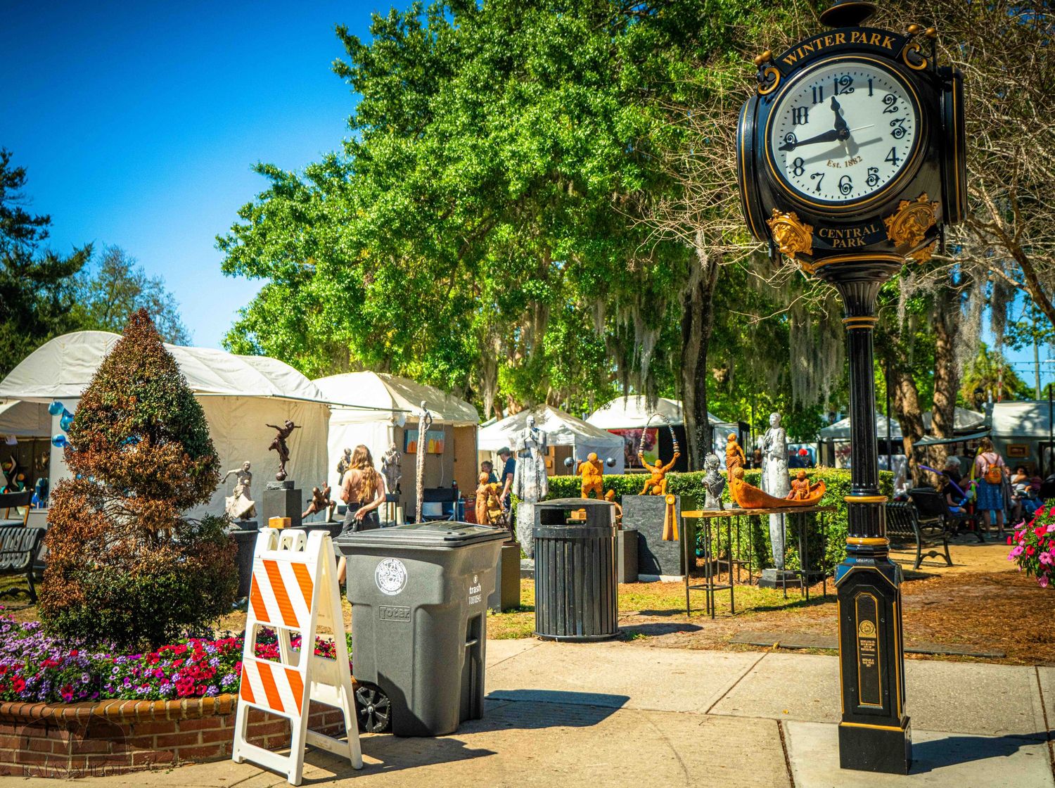 Outdoor festival marketplace with vendor tents, decorative clock post and landscaped walkway under blue skies.