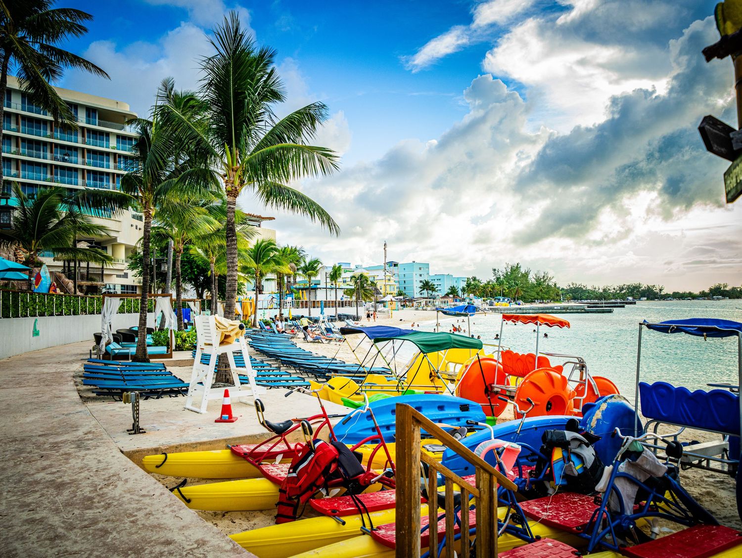 Colorful pedal boats and kayaks lined up on a tropical beach resort with palm trees and blue sky.