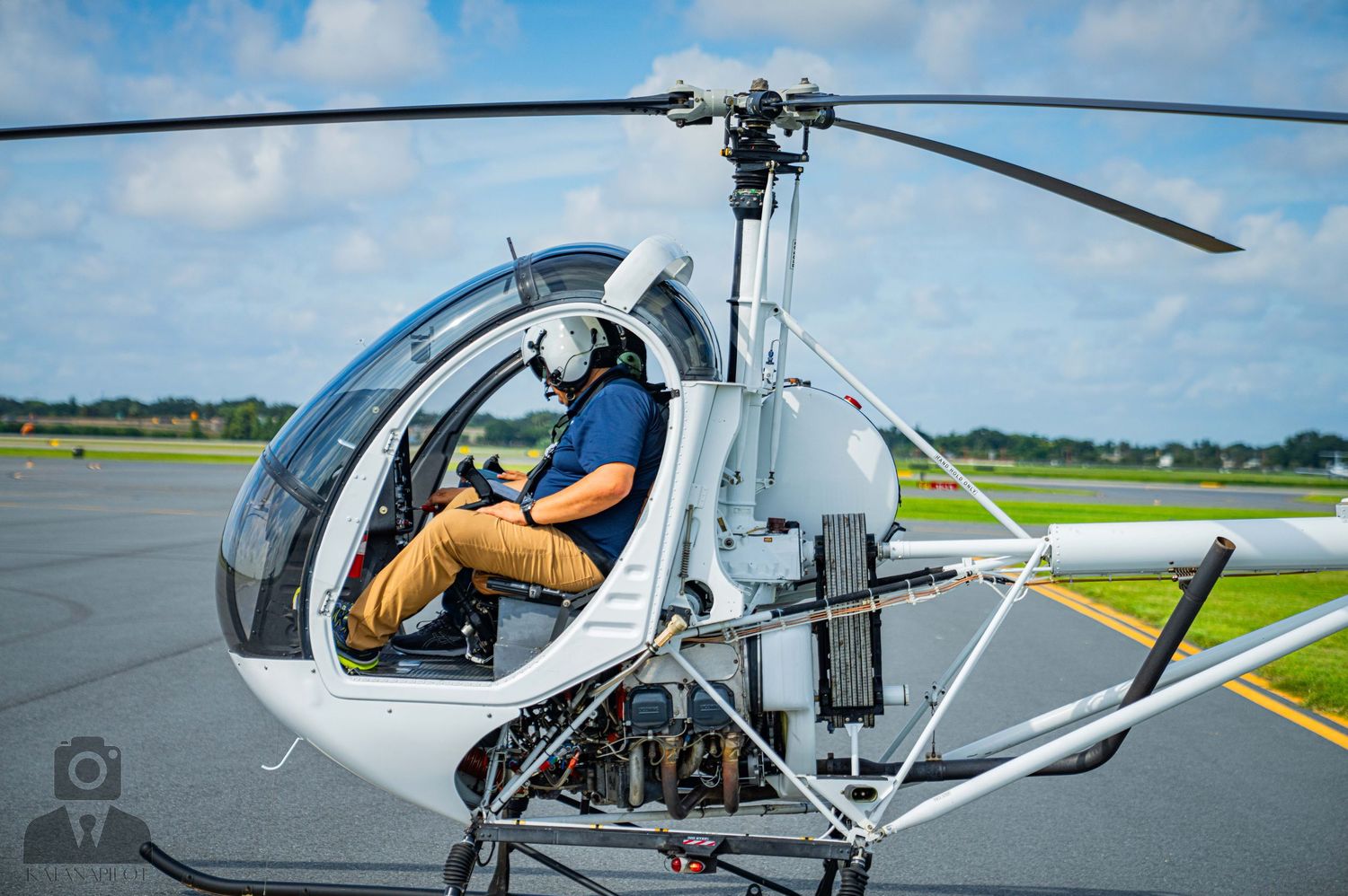 Pilot preparing for takeoff in small helicopter on airfield tarmac under blue skies.