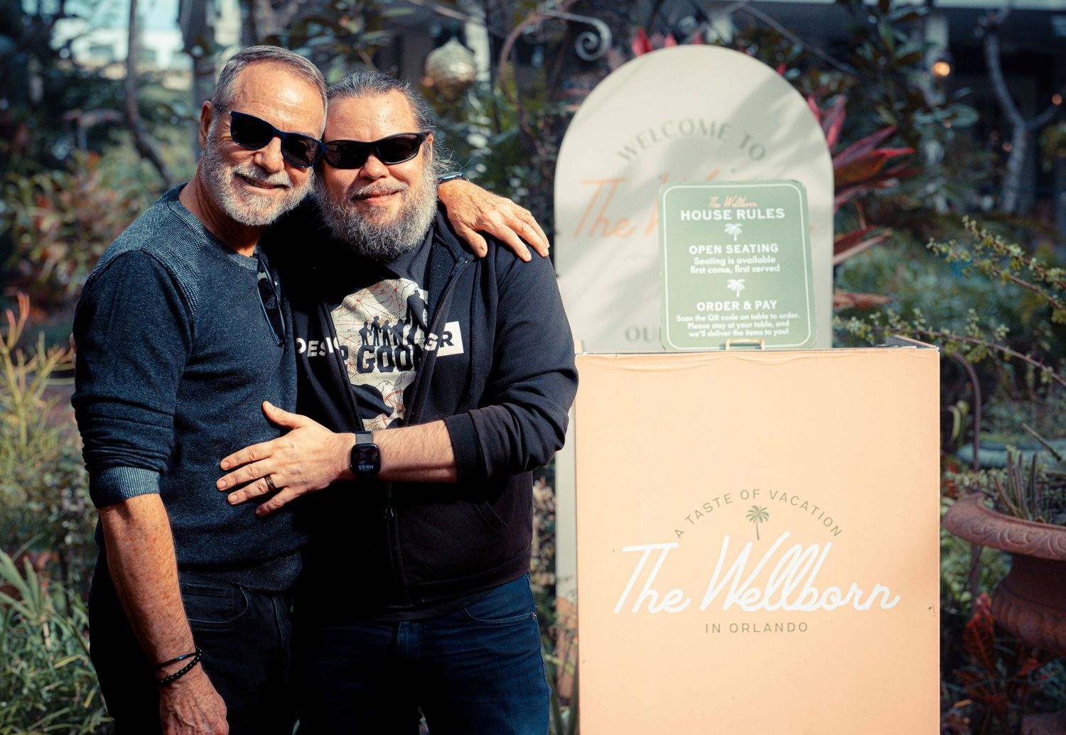 Two people in sunglasses embrace next to a cardboard box and decorative gravestone in an outdoor setting.