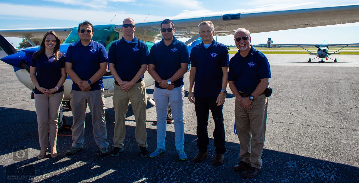 Group of aviation personnel standing together on tarmac with small aircraft in background.