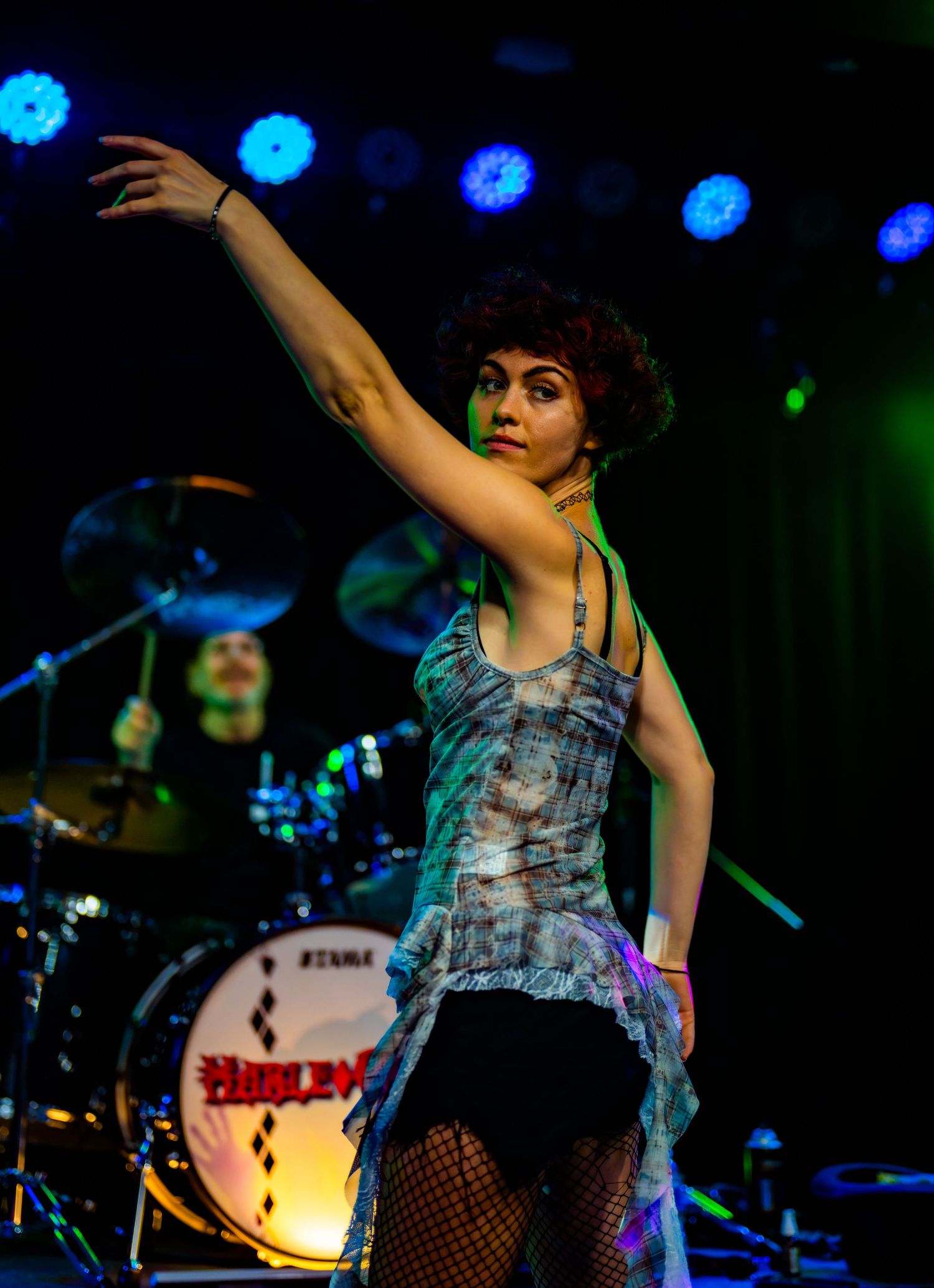 Female dancer performing on stage with arm raised, wearing fishnet stockings under colorful stage lights.