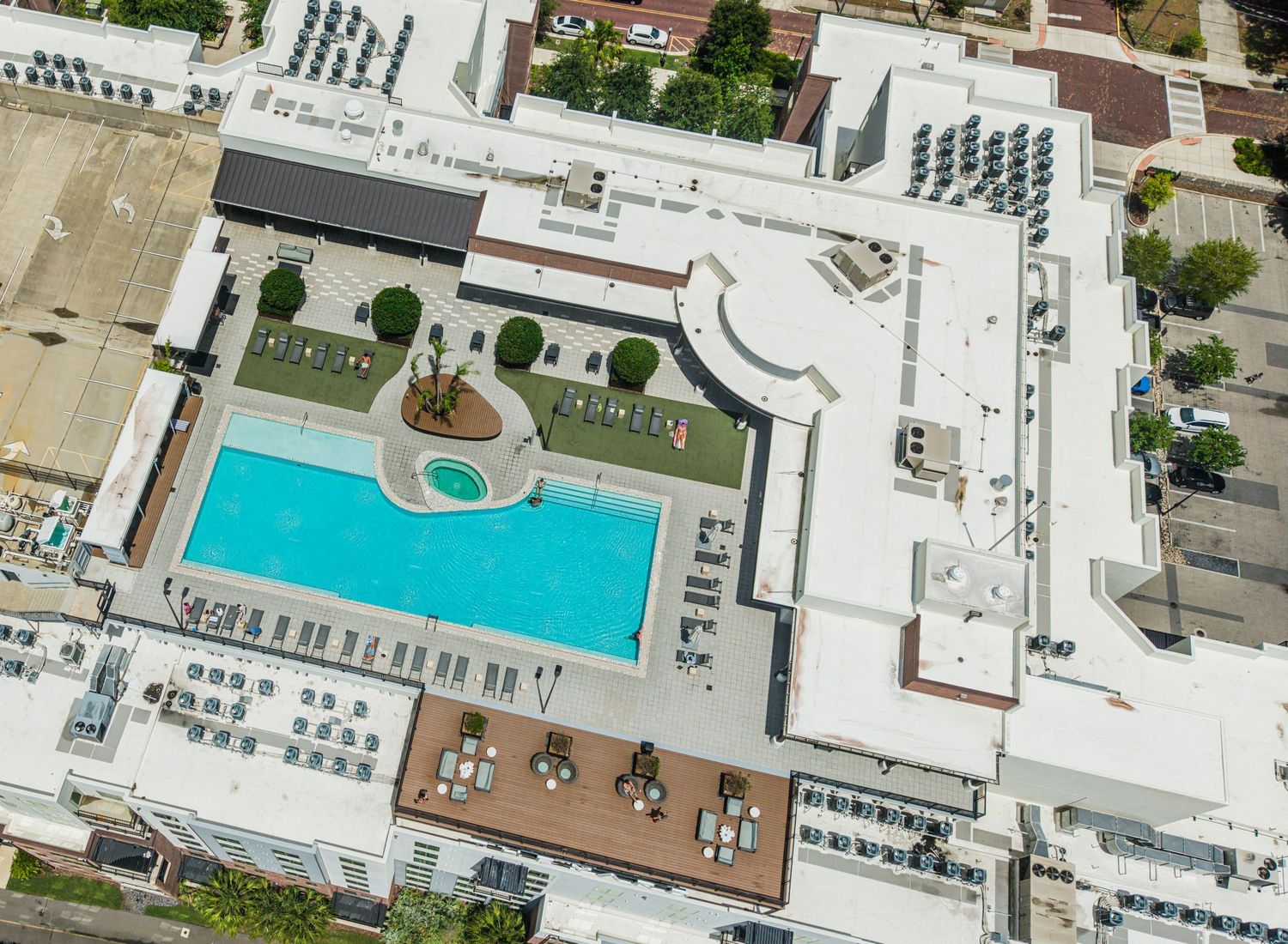 Aerial view of a luxury hotel rooftop pool surrounded by lounge chairs and manicured hedges in an urban setting.