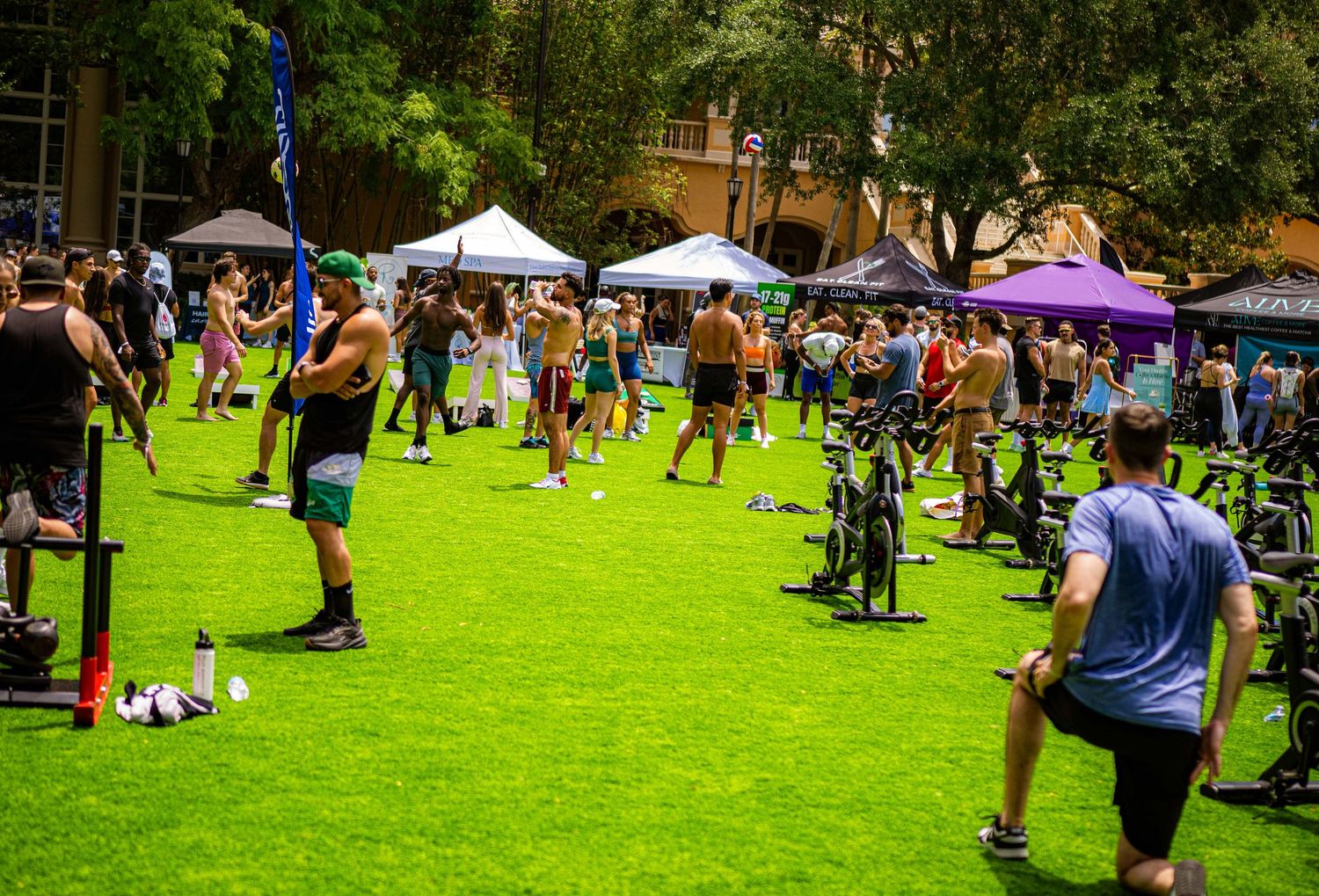 Outdoor fitness event on green turf with people exercising, cycling, and vendors under canopy tents on a sunny day.