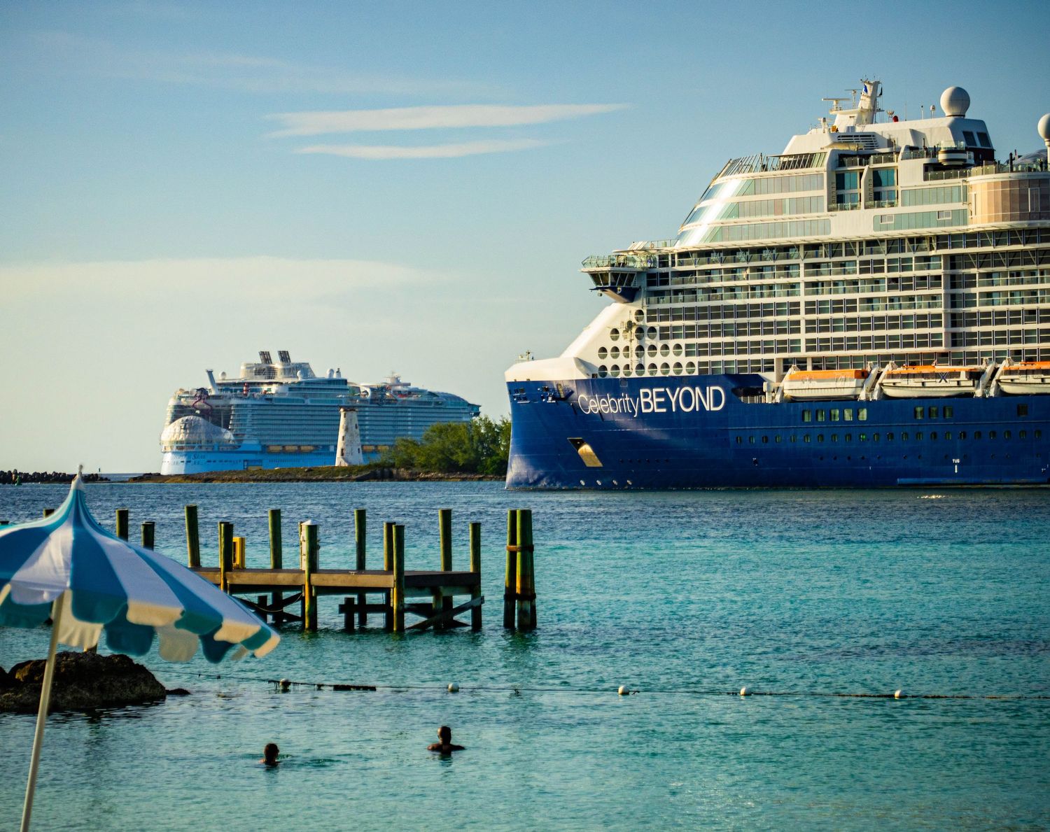 Celebrity Beyond cruise ship docked in turquoise Caribbean waters with a beach umbrella and wooden pier in the foreground.