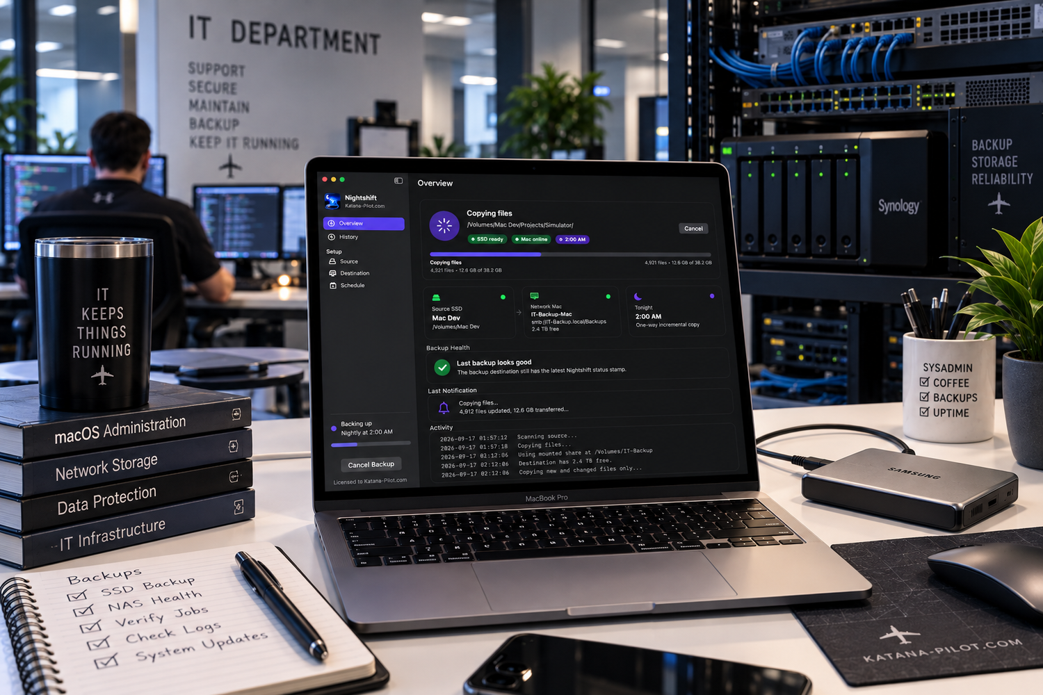IT department workspace with laptop displaying dark-themed software dashboard, networking books, and server racks in background.