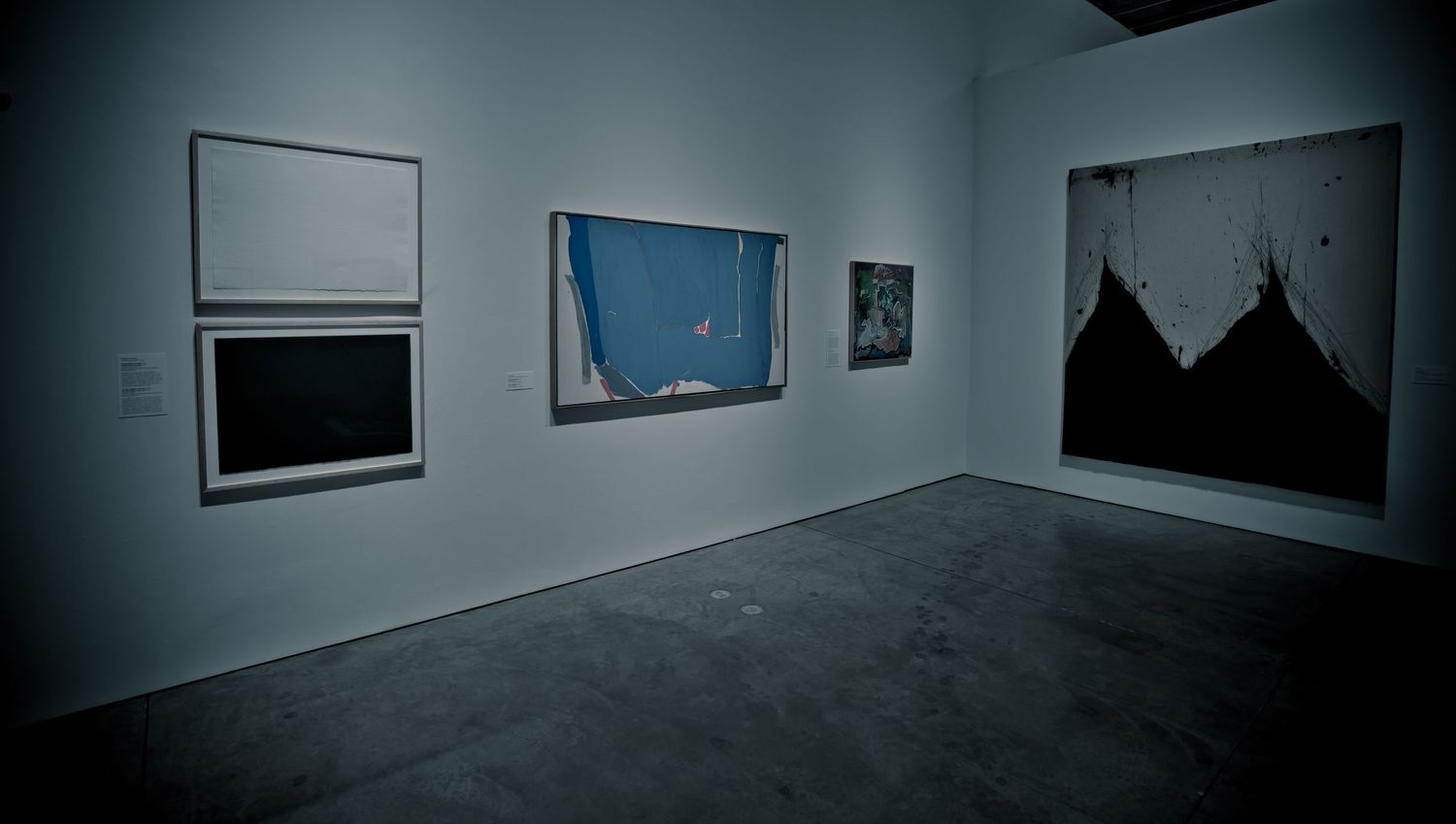 Art gallery interior displaying three abstract paintings on white walls with dark concrete floors in dim lighting.