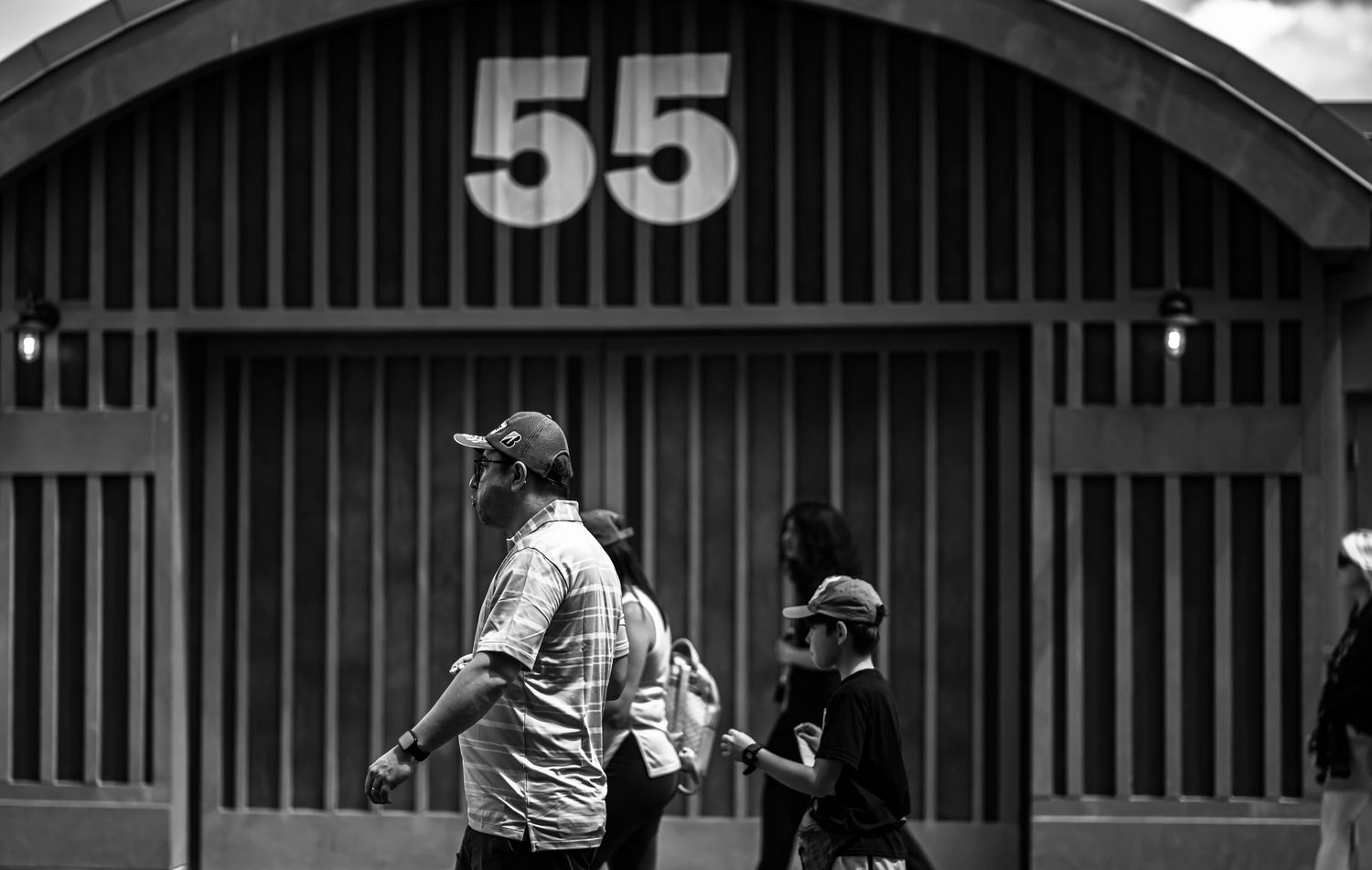A black and white photo shows people walking past numbered warehouse doors and buildings in an urban setting.