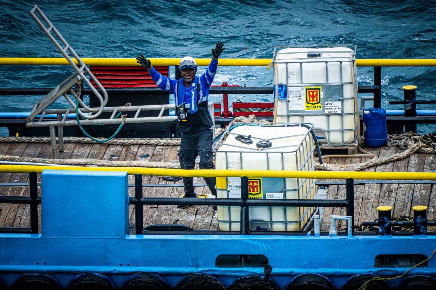 Worker in blue jacket waves from a industrial barge deck with cargo containers on choppy ocean waters.