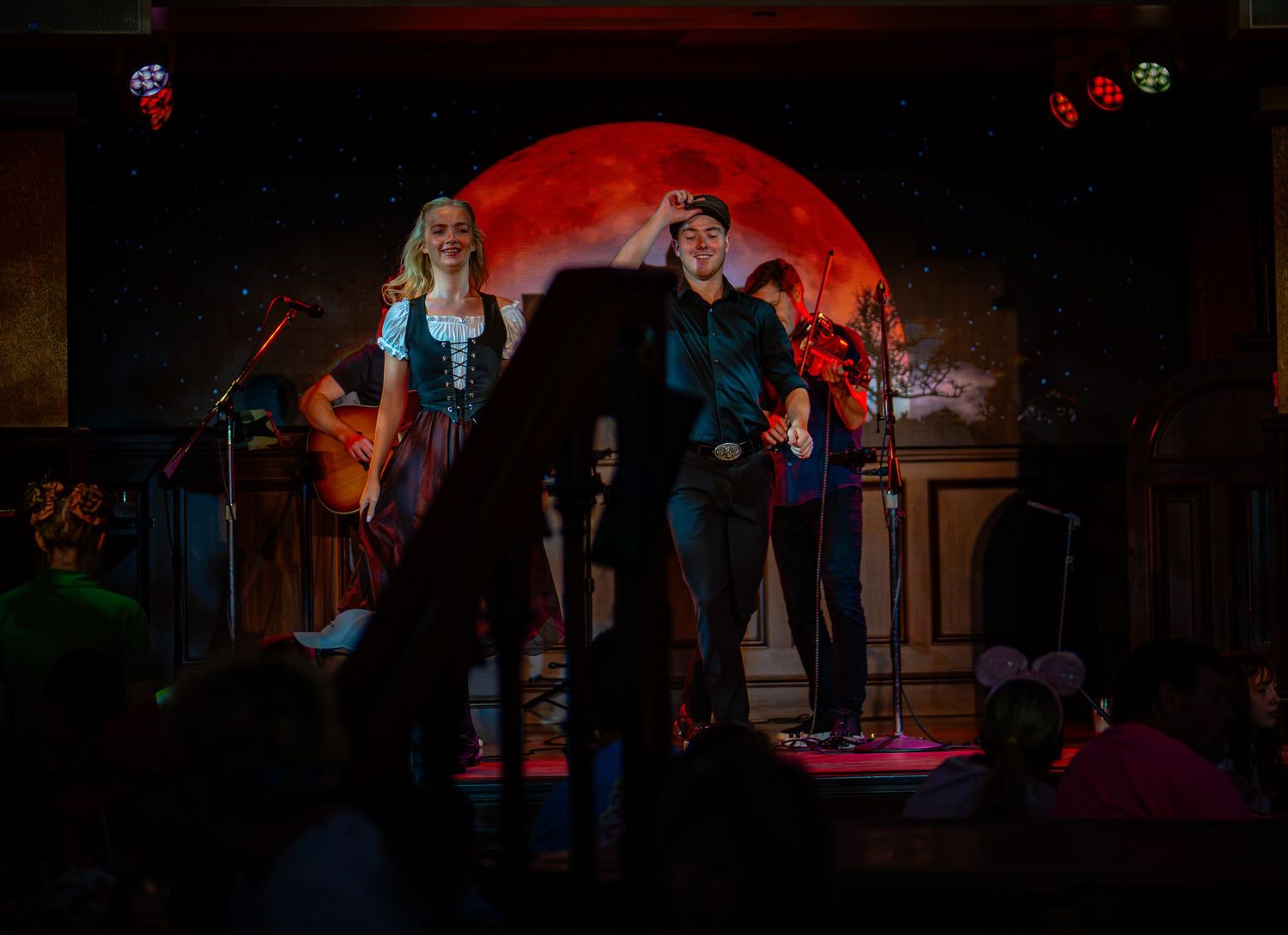 Two performers on stage with a glowing red moon backdrop in a dimly lit venue.