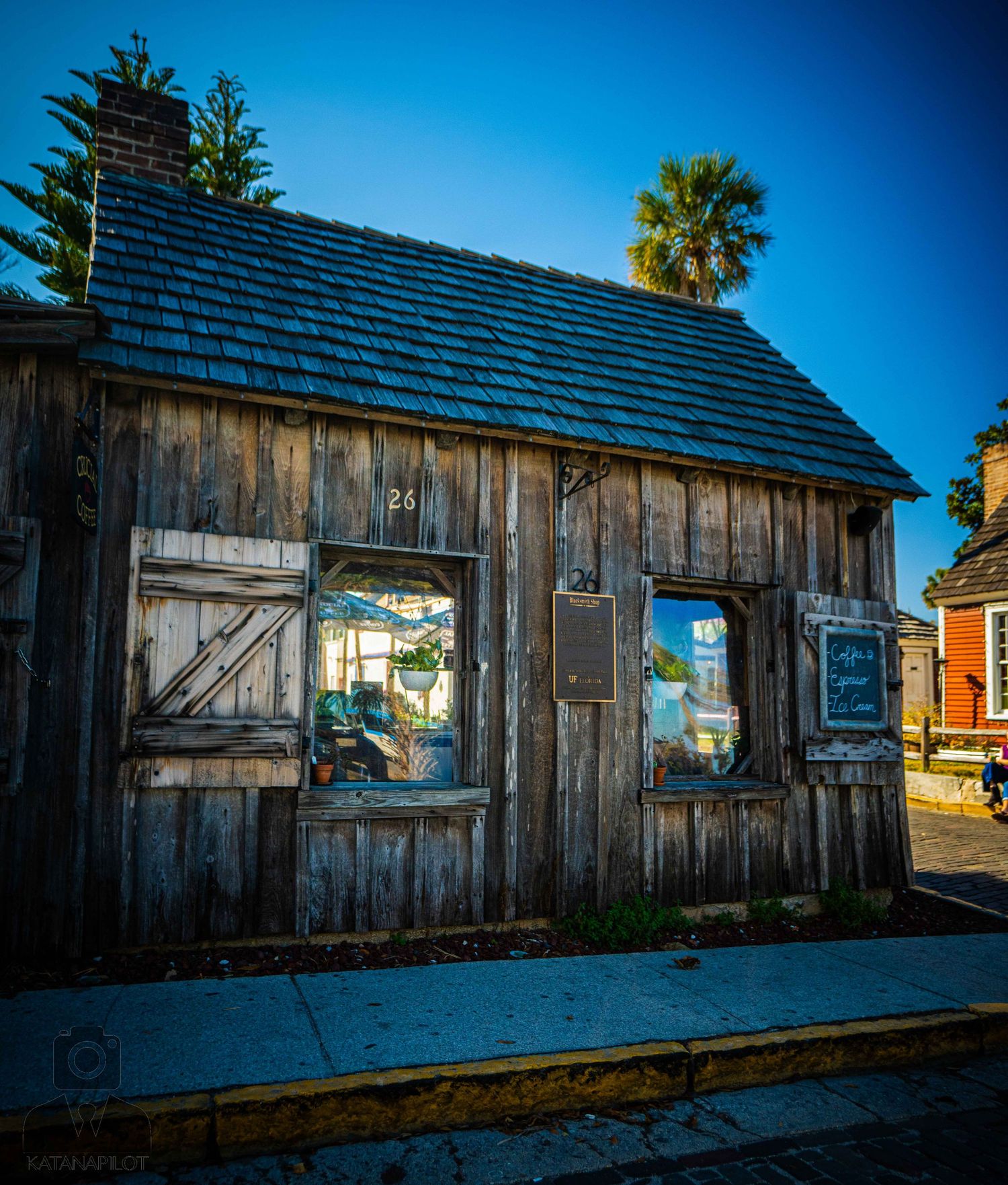 Rustic wooden cabin with weathered boards and shingled roof against bright blue sky with palm trees in background.