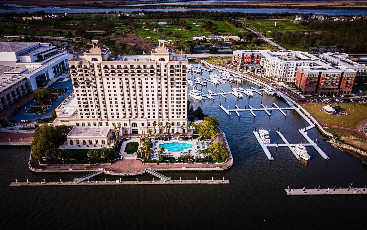 Aerial view of waterfront hotels and marinas with docked boats along a coastal shoreline at sunset.