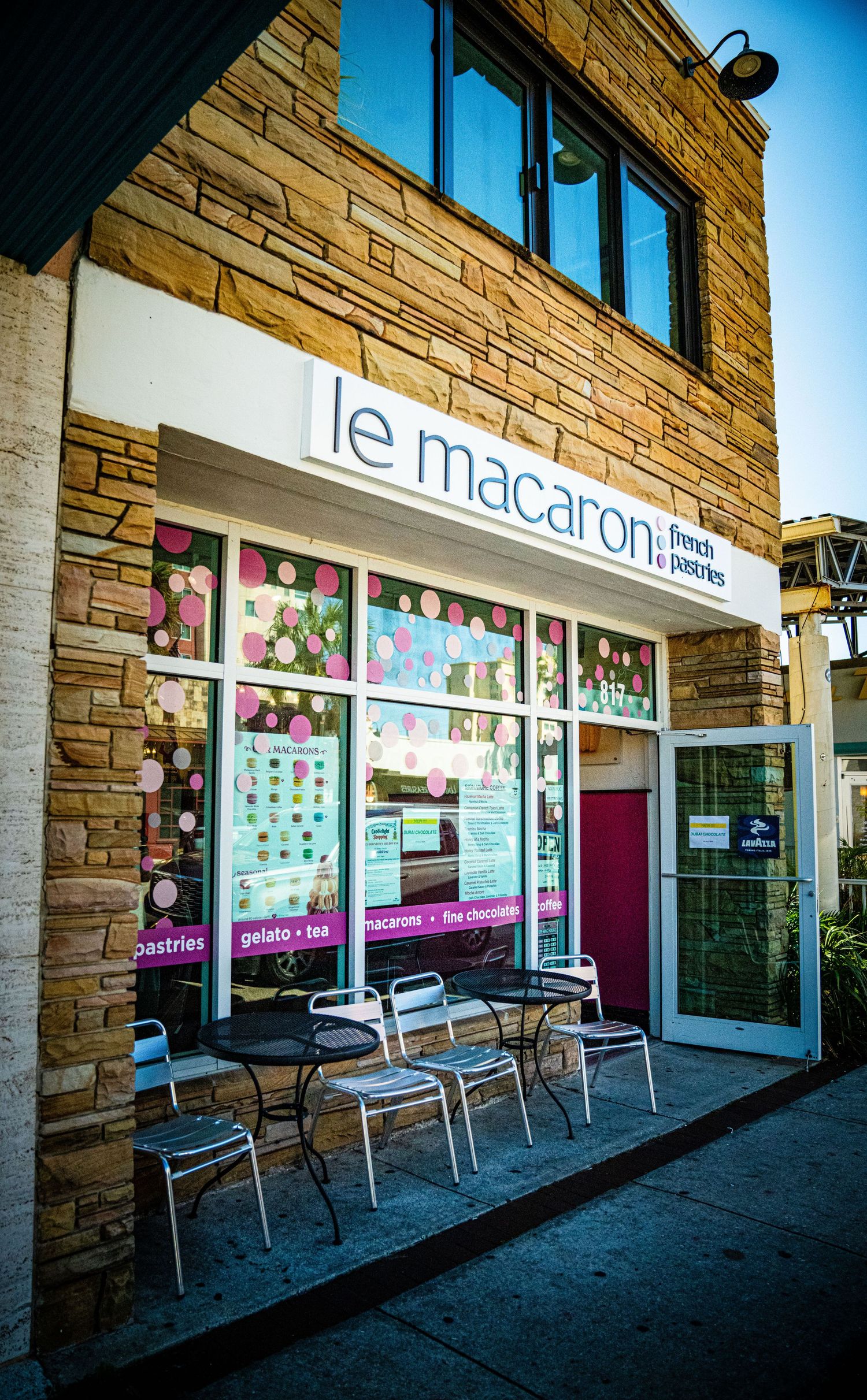 Le Macaron French pastry shop storefront with outdoor seating and stone facade in a shopping center.
