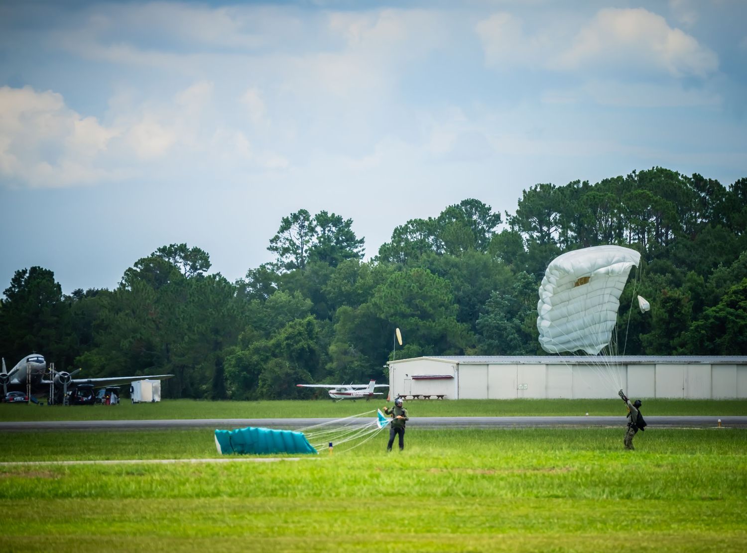 Wide shot of skydivers landing on green field near industrial buildings.