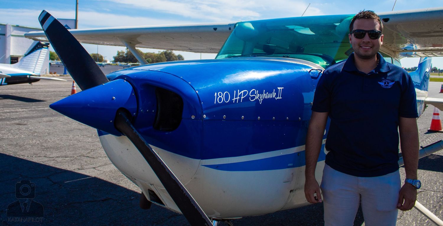 Blue and white private aircraft parked on runway with pilot standing beside.