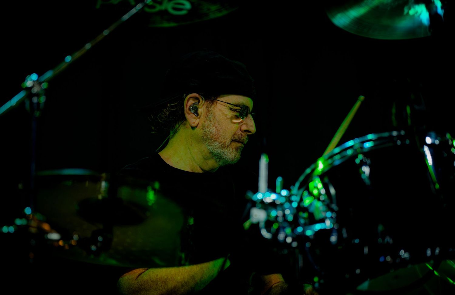 Drummer performing on stage in dark lighting with green accents, focused behind a drum kit.