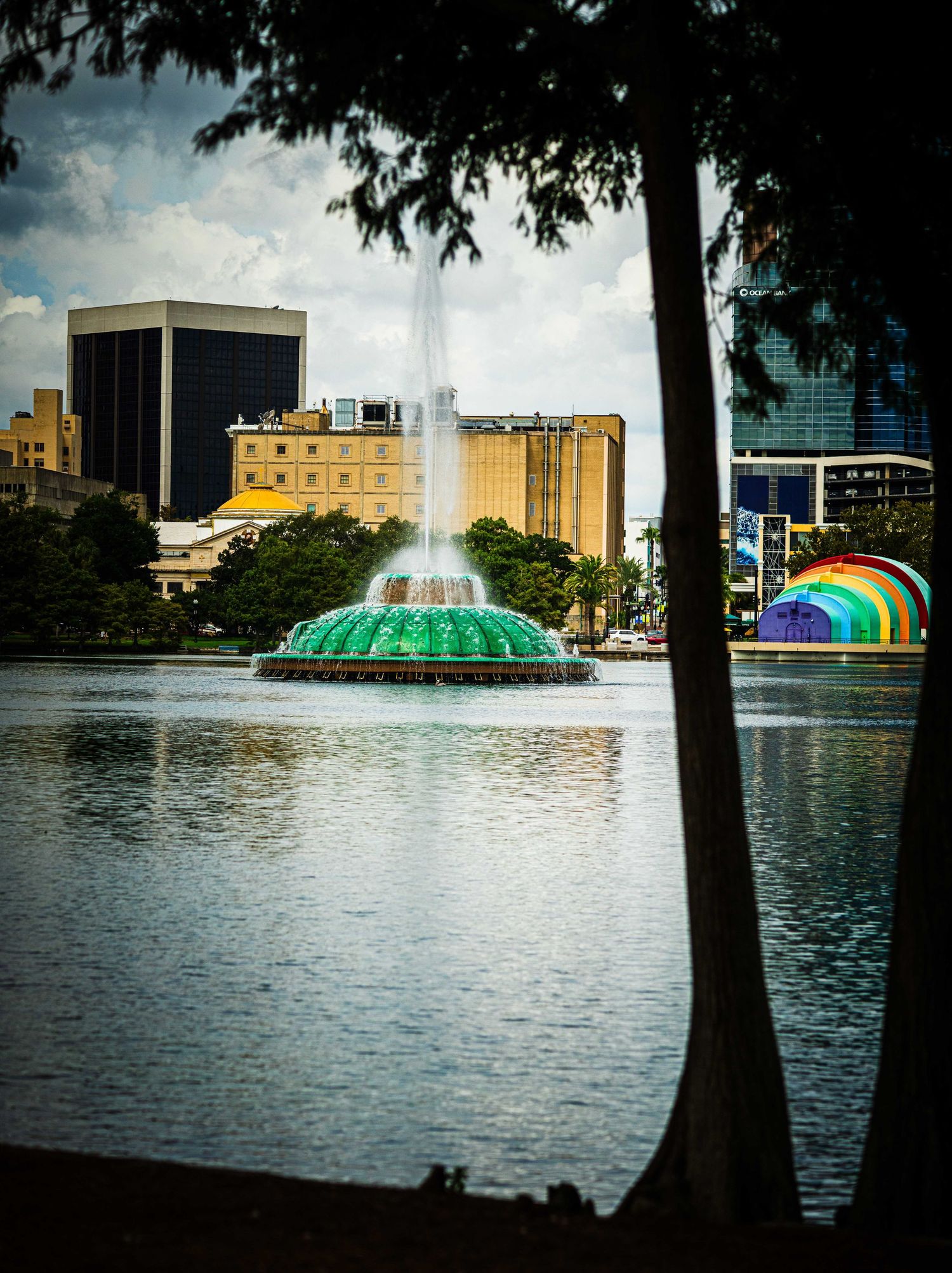 Green fountain jets water skyward on Lake Eola in Orlando, FL, with city skyline and rainbow amphitheater visible.