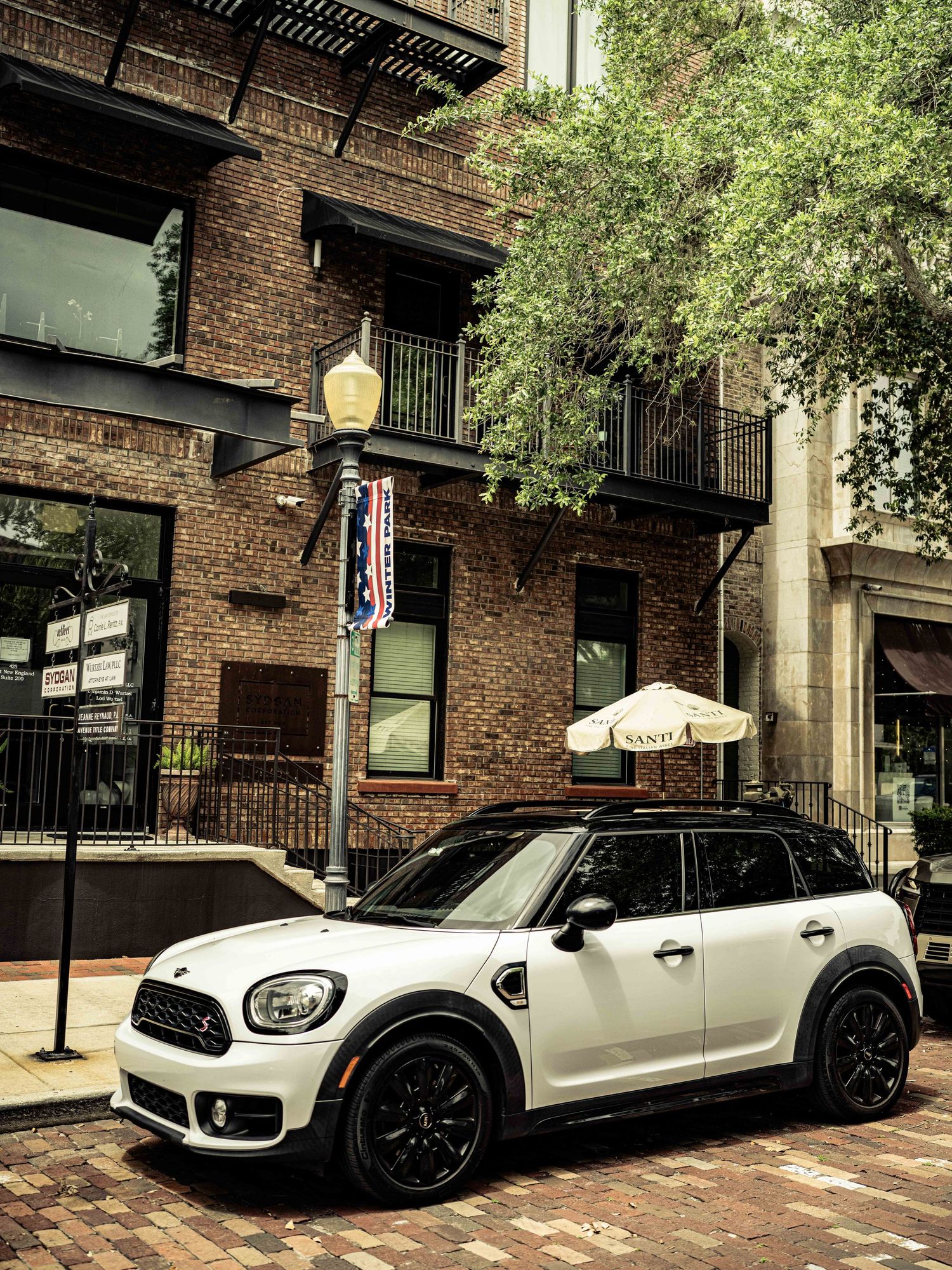 White MINI Cooper Countryman parked on brick street beside lamppost with historic brick buildings and trees in urban setting.