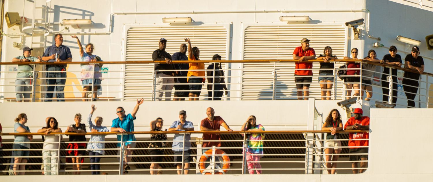 Passengers line the railings of a cruise ship deck as they look out over the water and port below.