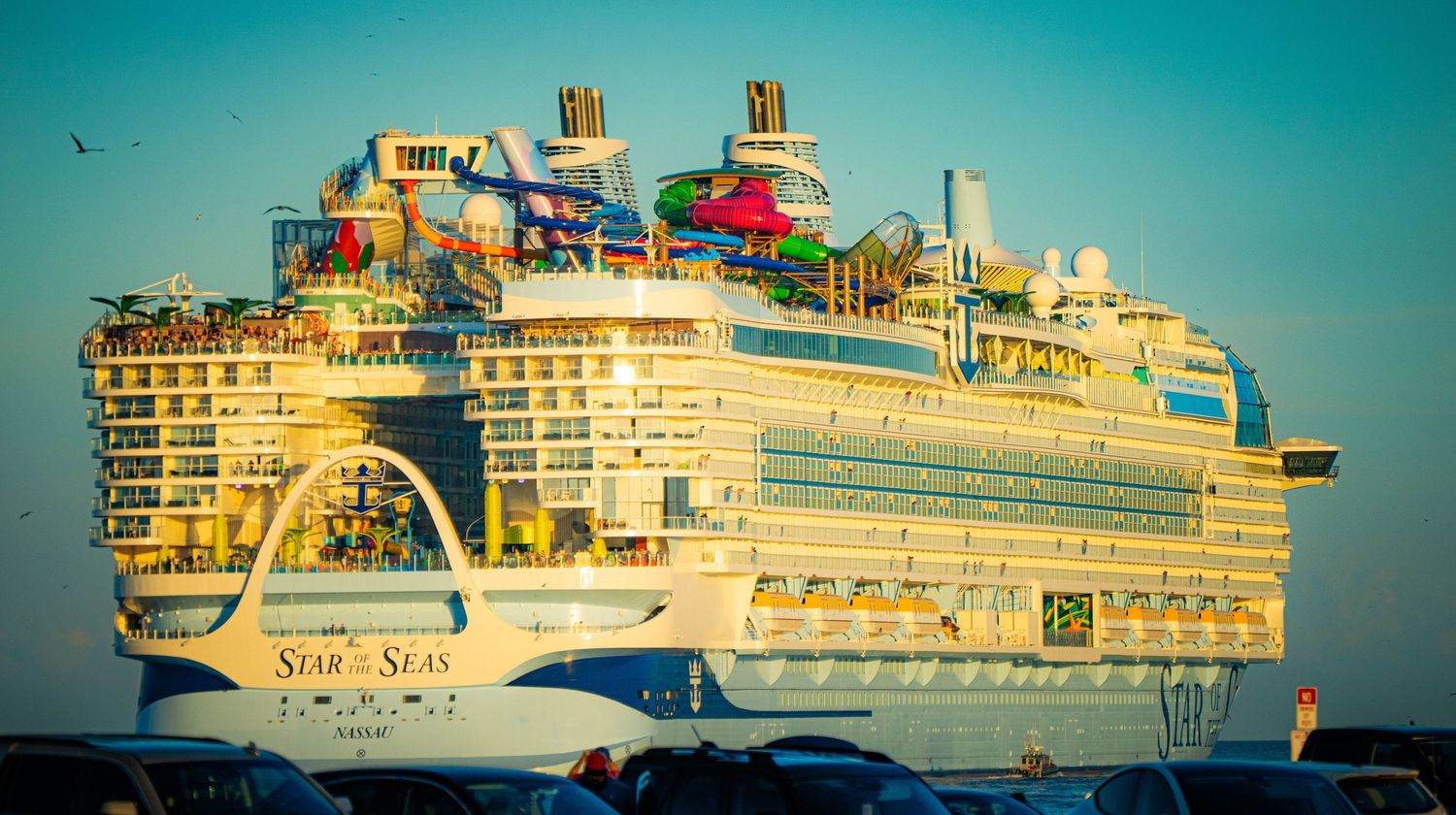 A massive Royal Caribbean cruise ship with colorful water slides and amenities glows in golden sunlight against a turquoise sky.