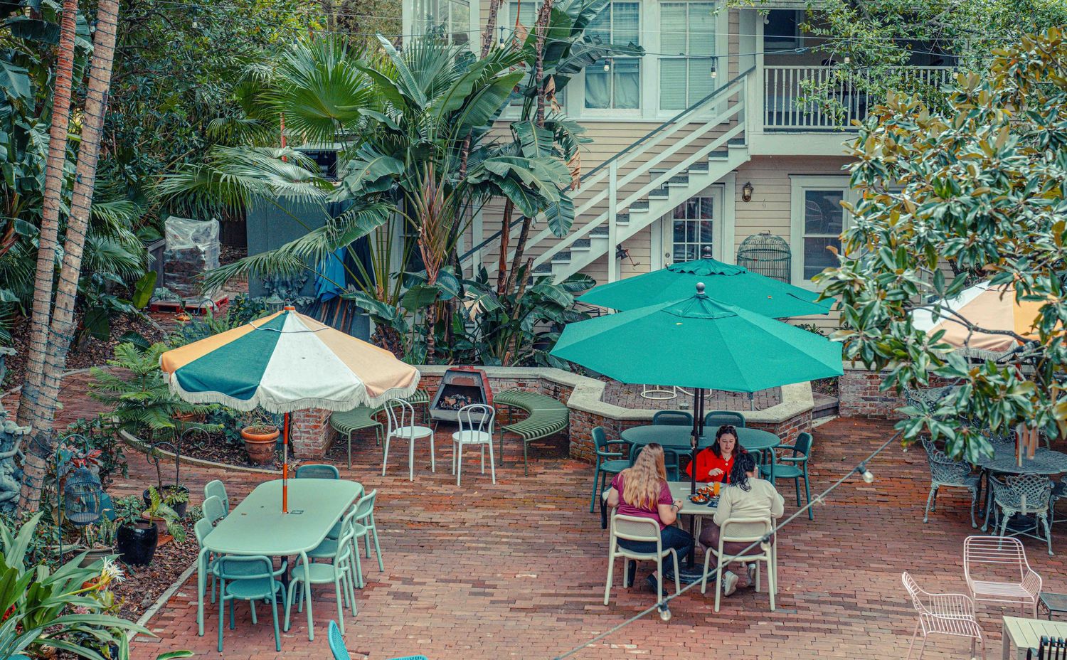 Outdoor patio dining area with green umbrellas and metal chairs surrounded by lush tropical plants and palm trees.