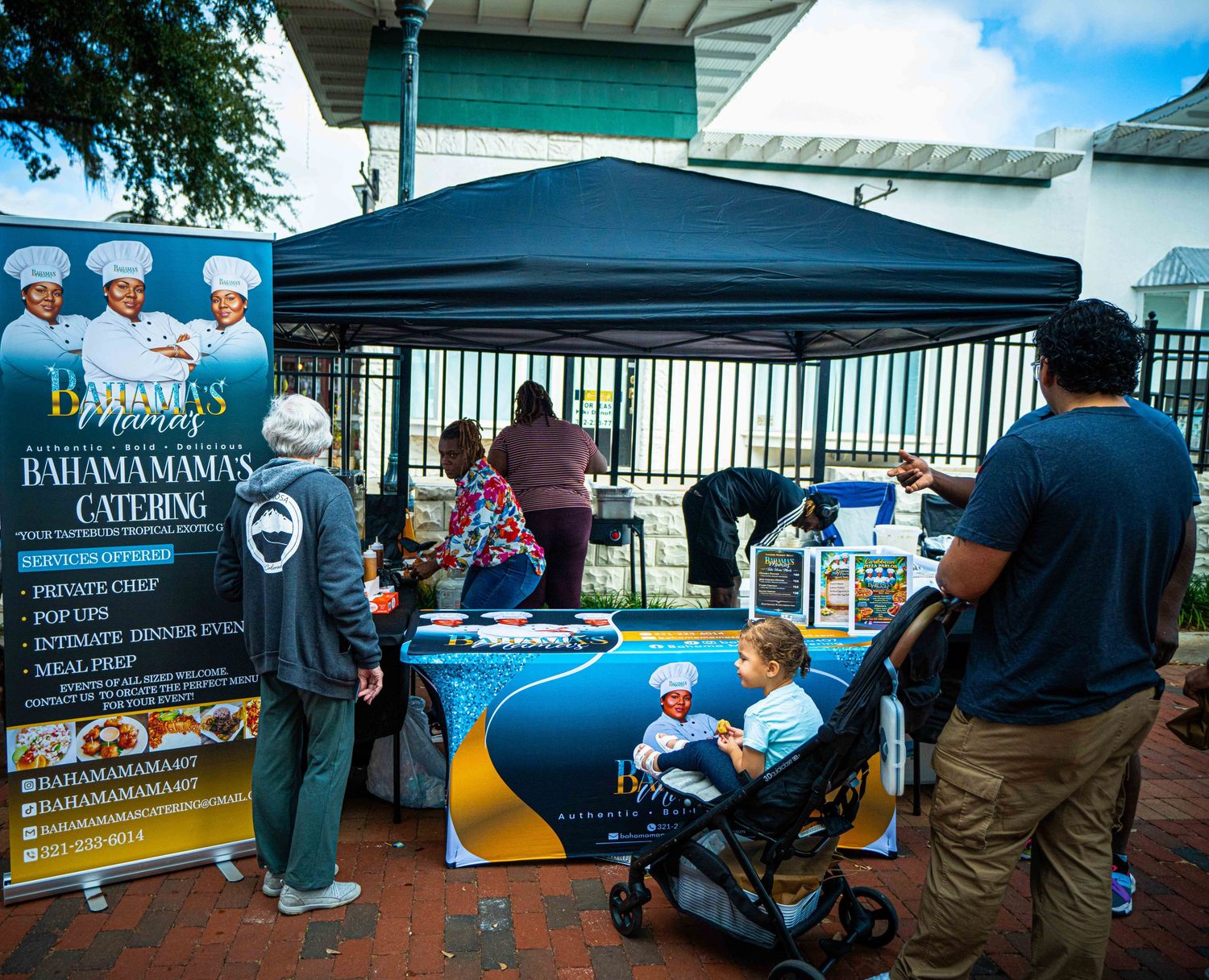 Outdoor catering setup with blue tent, banner stand, and customers at a food service station.
