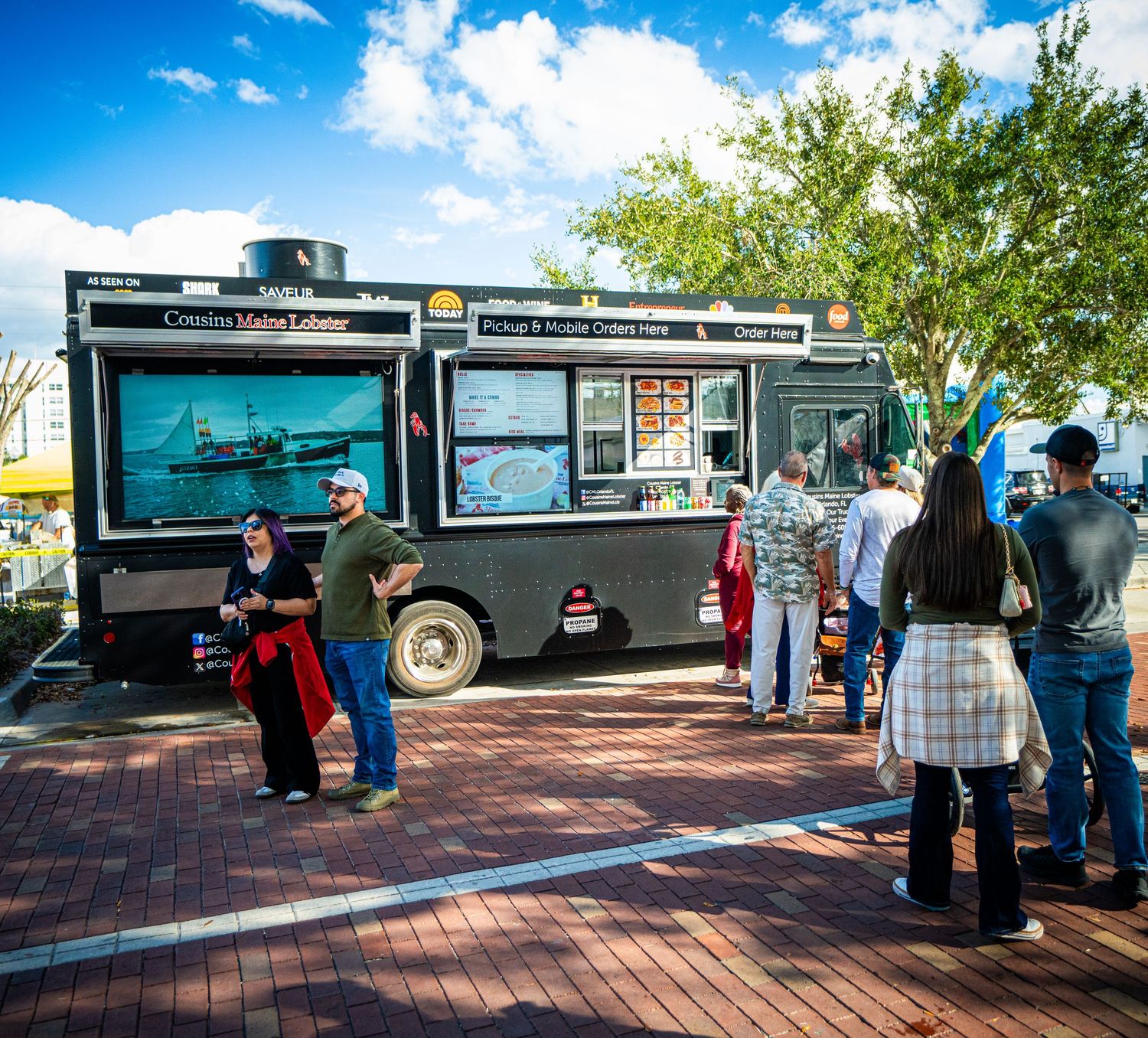 Food trucks line up along a brick paved walkway under blue skies during a sunny outdoor event.