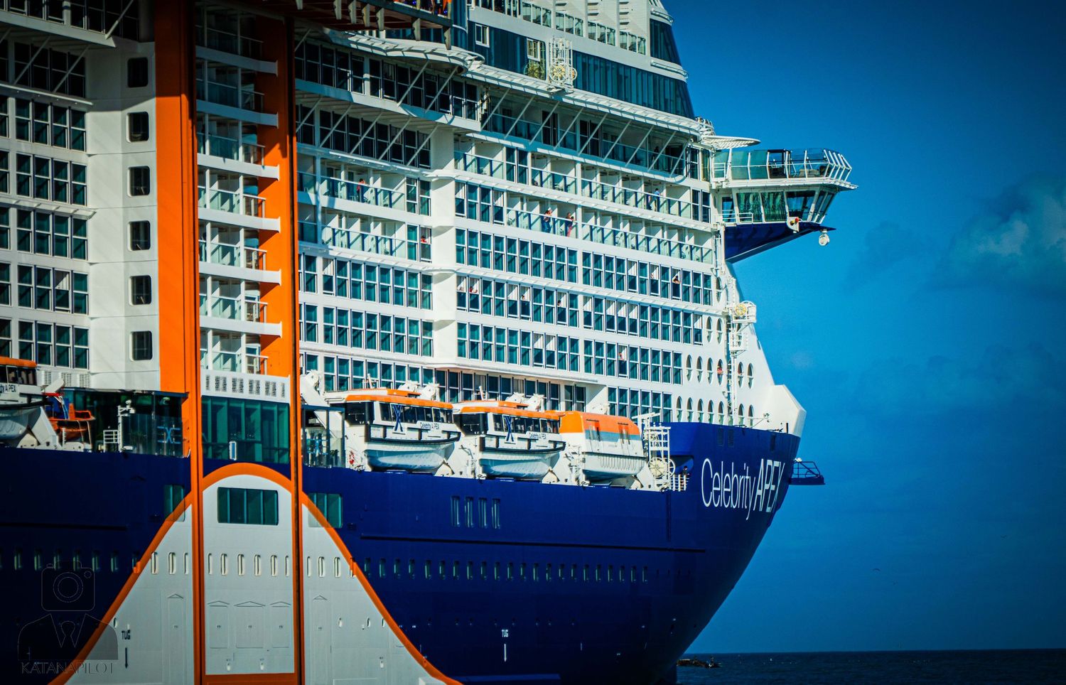 Luxury cruise ship with white and blue exterior docked at port against bright blue sky.