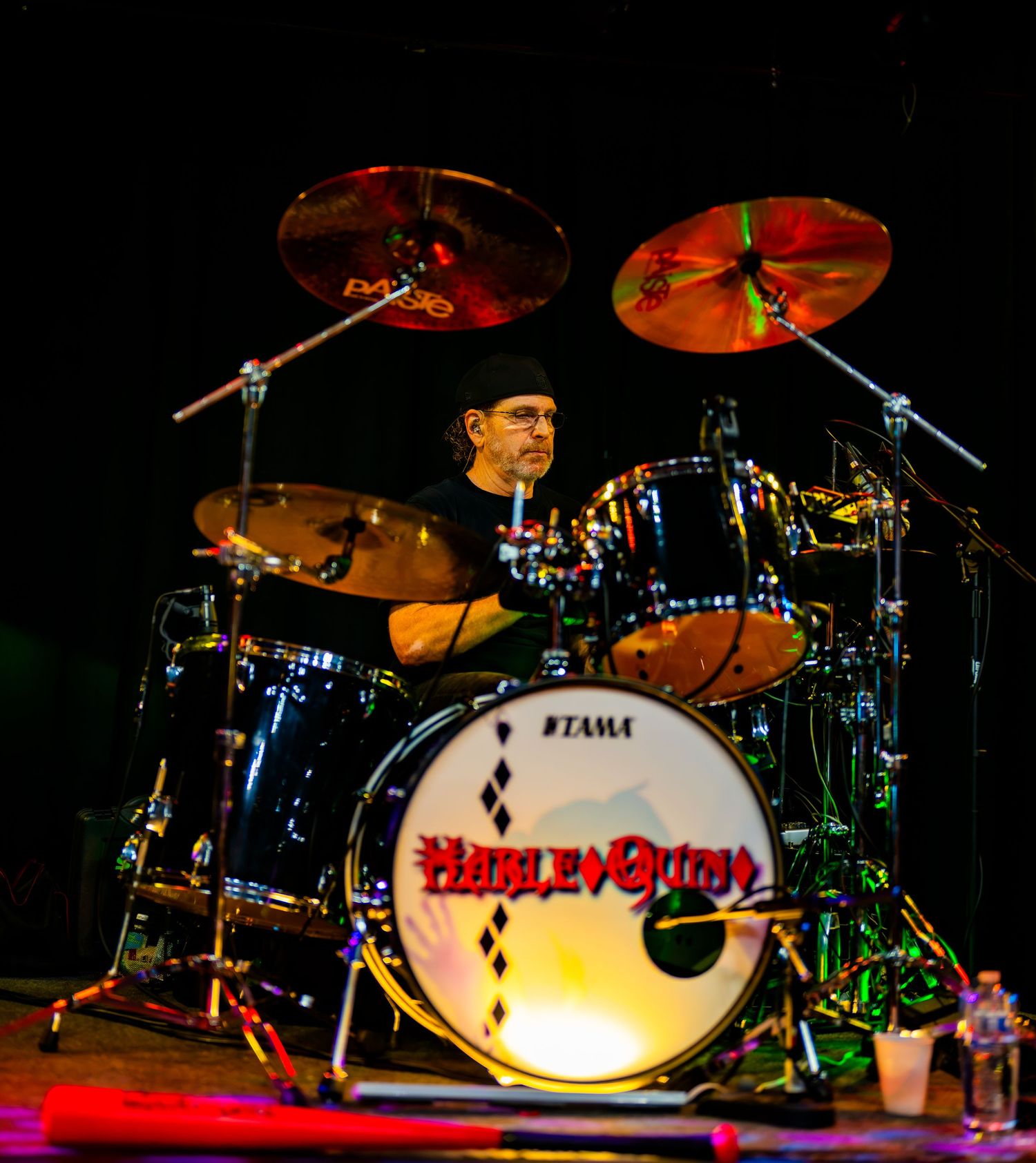 Drummer performing on a Tama drum kit with Harley Quinn branding under red stage lights.