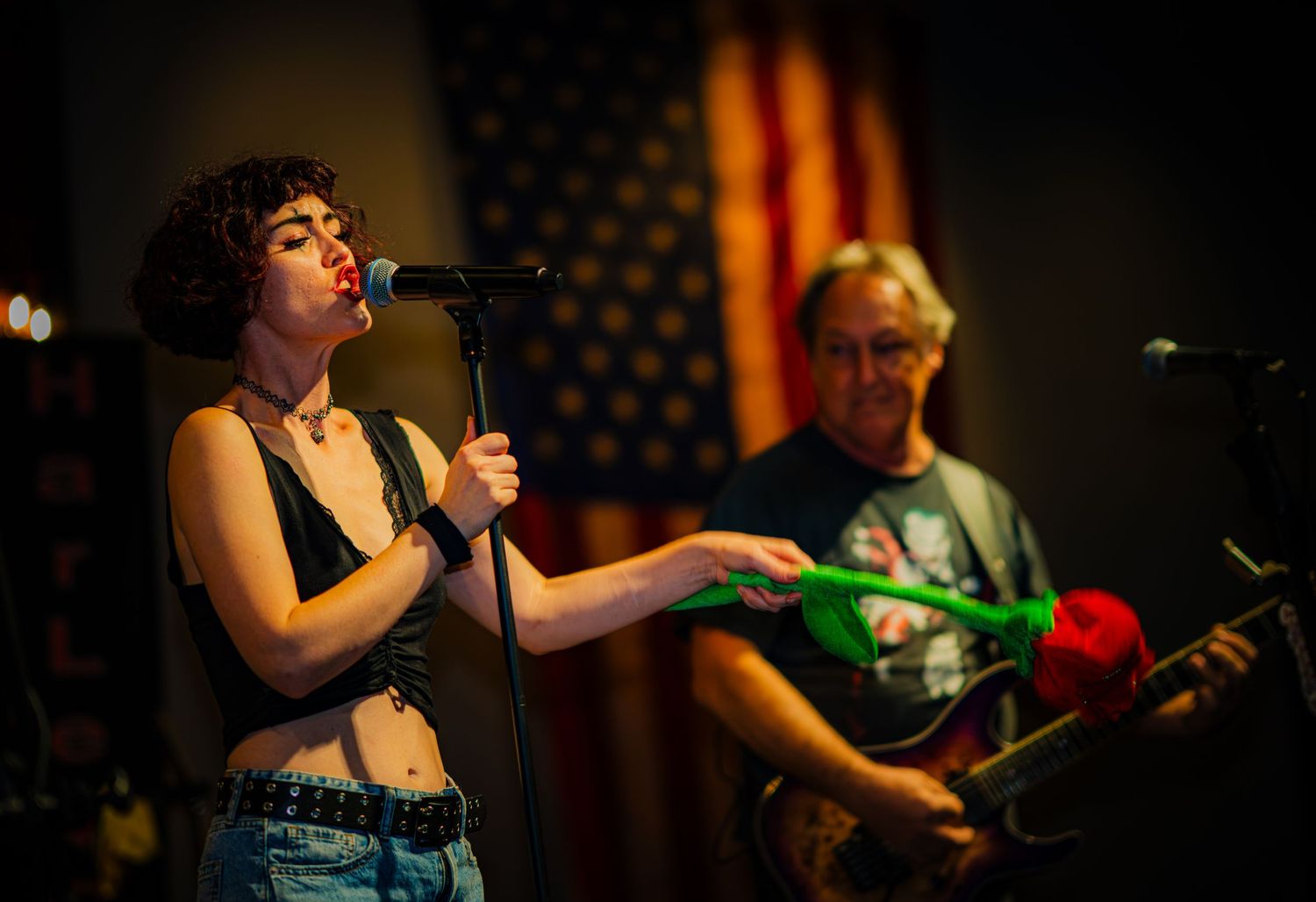 Singer performs with guitarist against American flag backdrop at music venue.