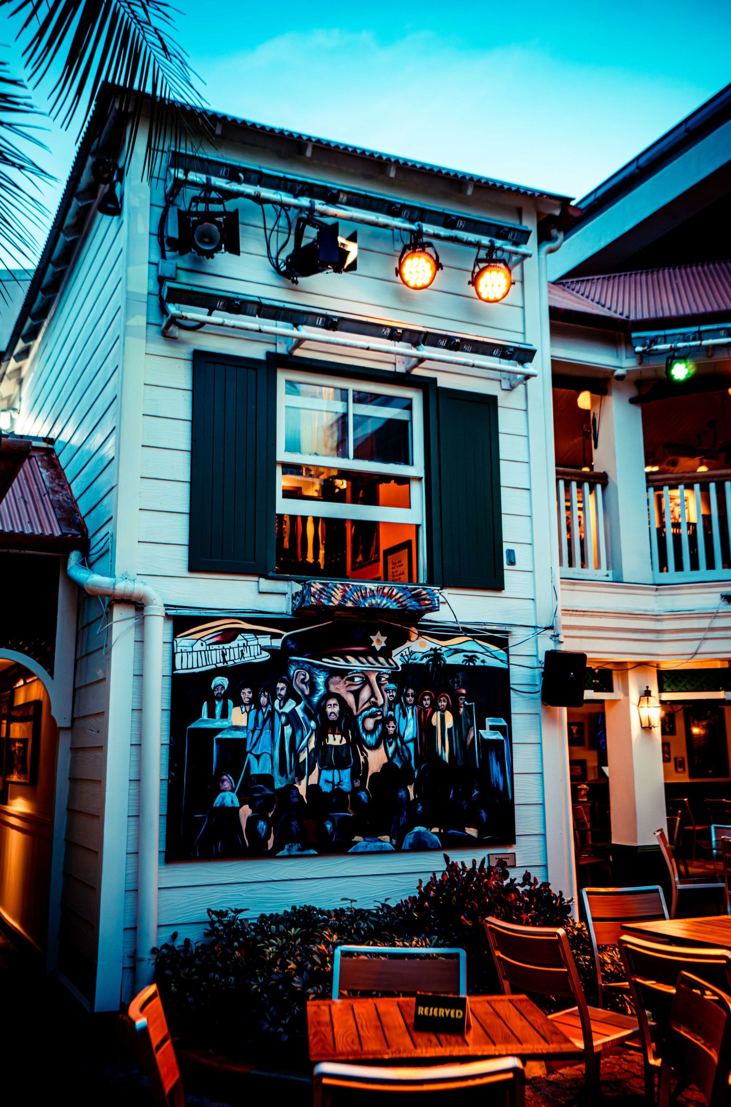 A modern white building with outdoor seating and umbrellas illuminated at dusk with a tropical ambiance.