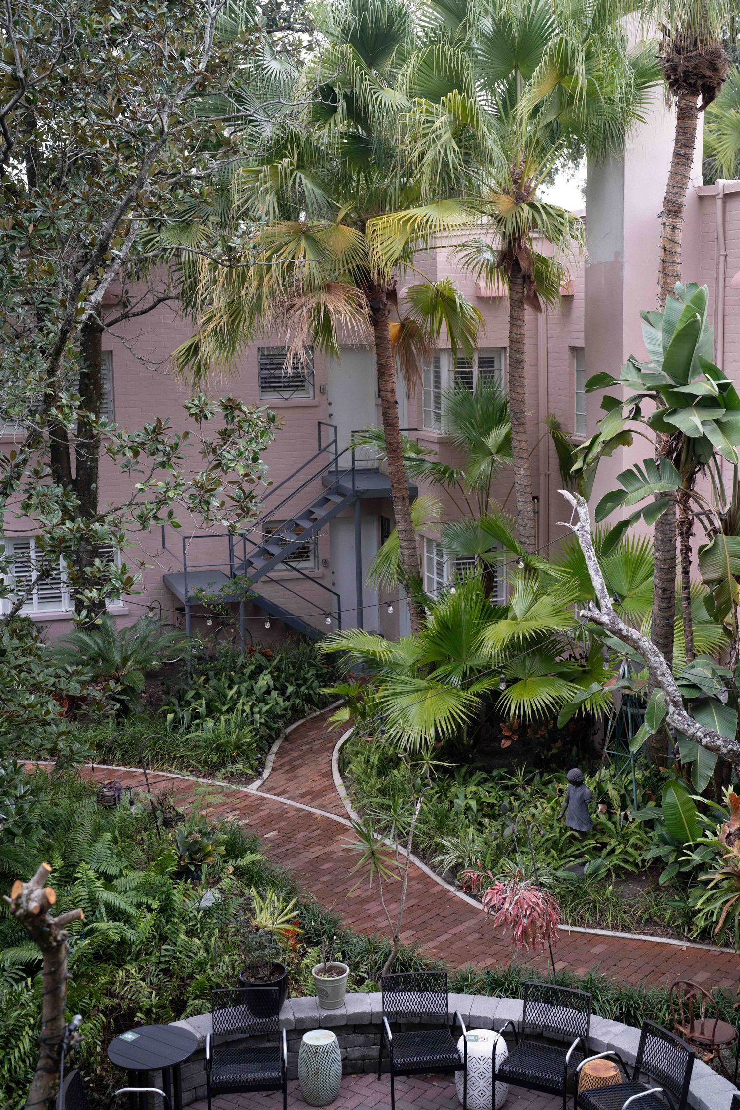Tropical courtyard garden with palm trees, lush greenery, and pink stucco buildings in the background.