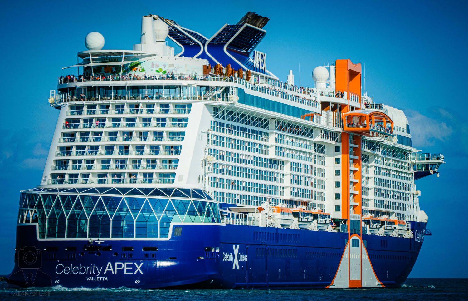 Celebrity Apex cruise ship with distinctive blue and orange funnel against bright blue sky.