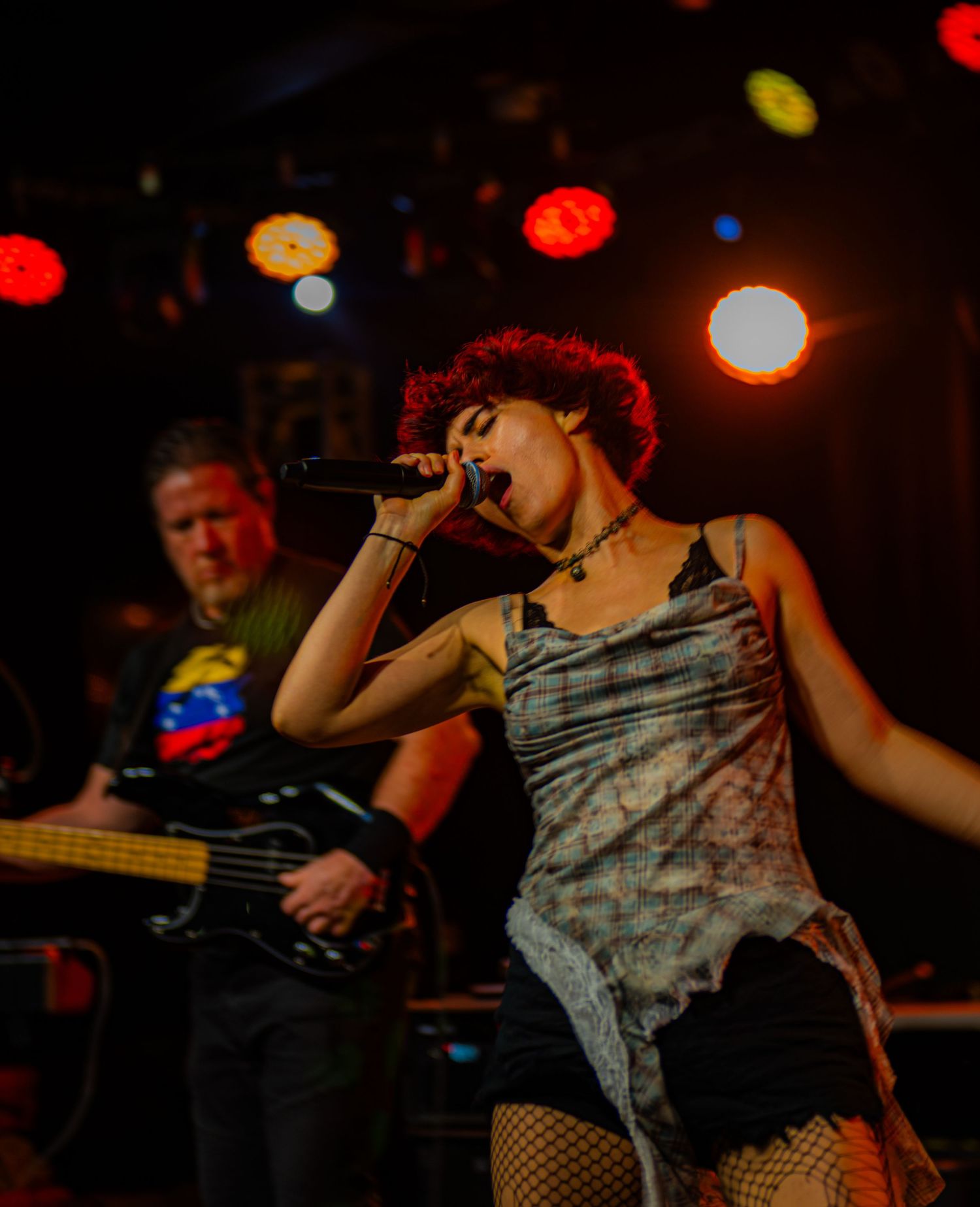 Female vocalist with red hair sings passionately on stage while guitarist performs behind her at live concert.