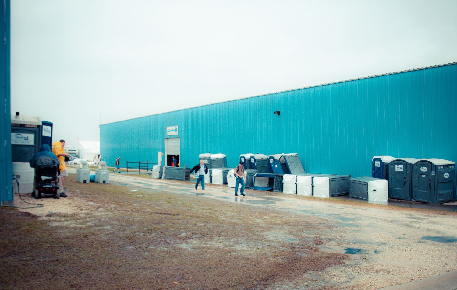Workers gather near loading docks and dumpsters along a large turquoise industrial warehouse building.