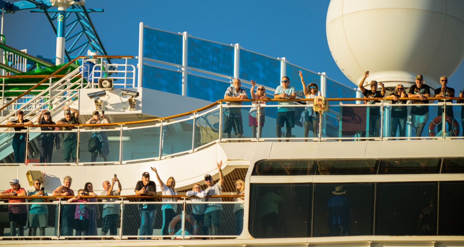 Cruise ship passengers gather on multiple decks near a large white radar dome during a sunny day at sea.