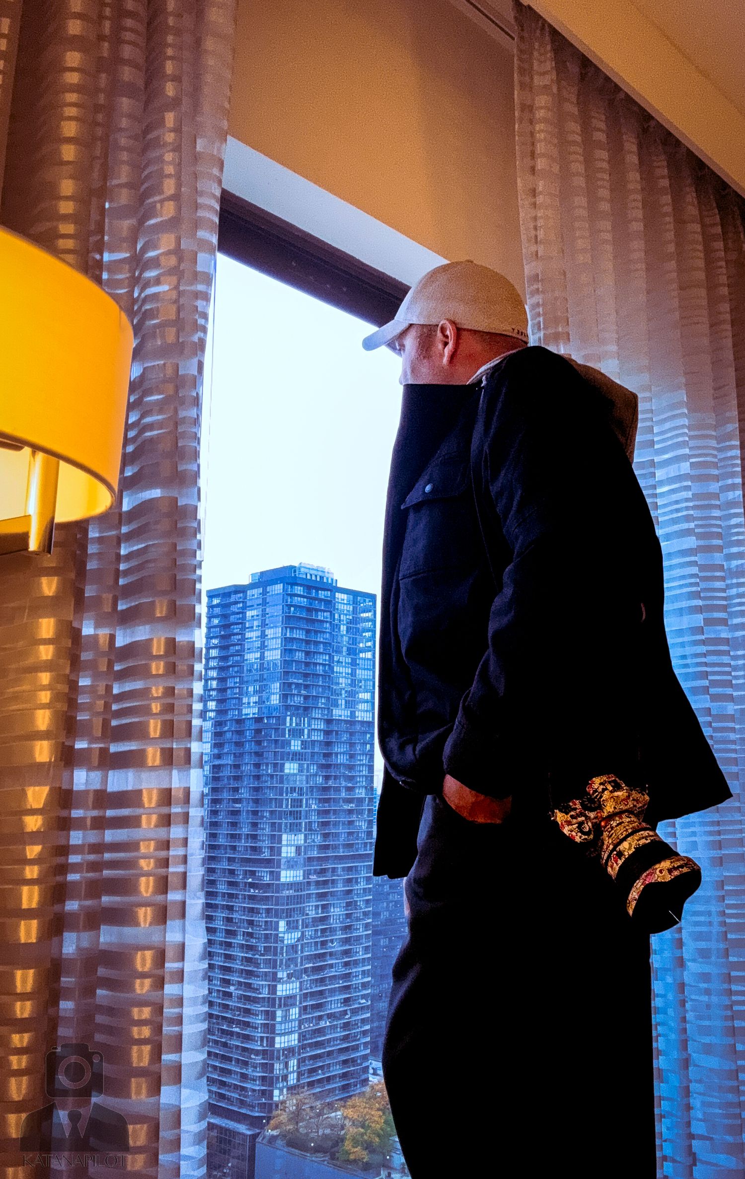 Silhouetted figure stands by window with city skyline and curtains in warm evening light.