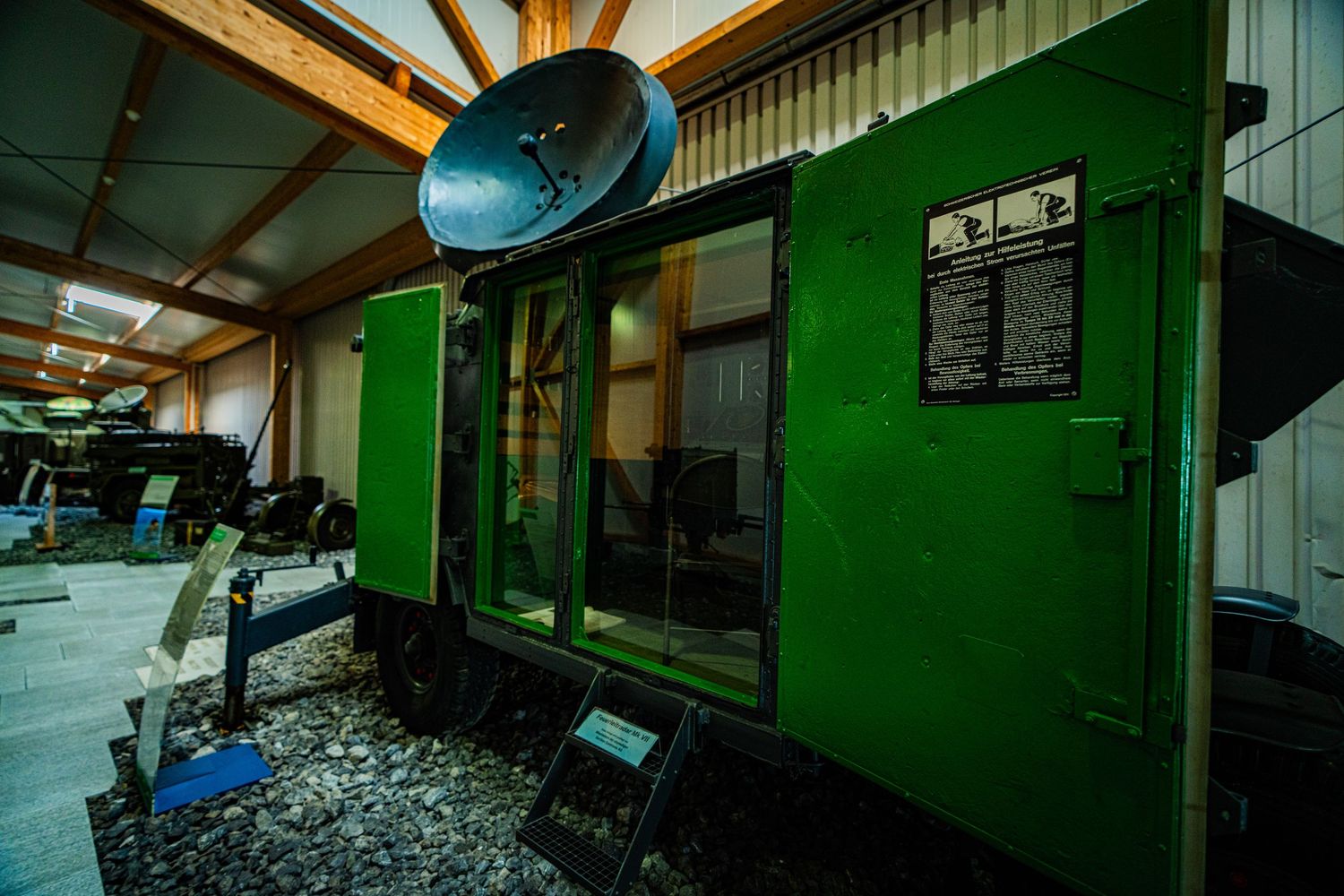 Green vintage military equipment shelter with a satellite dish mounted on top, displayed inside a museum hall with an informational plaque.