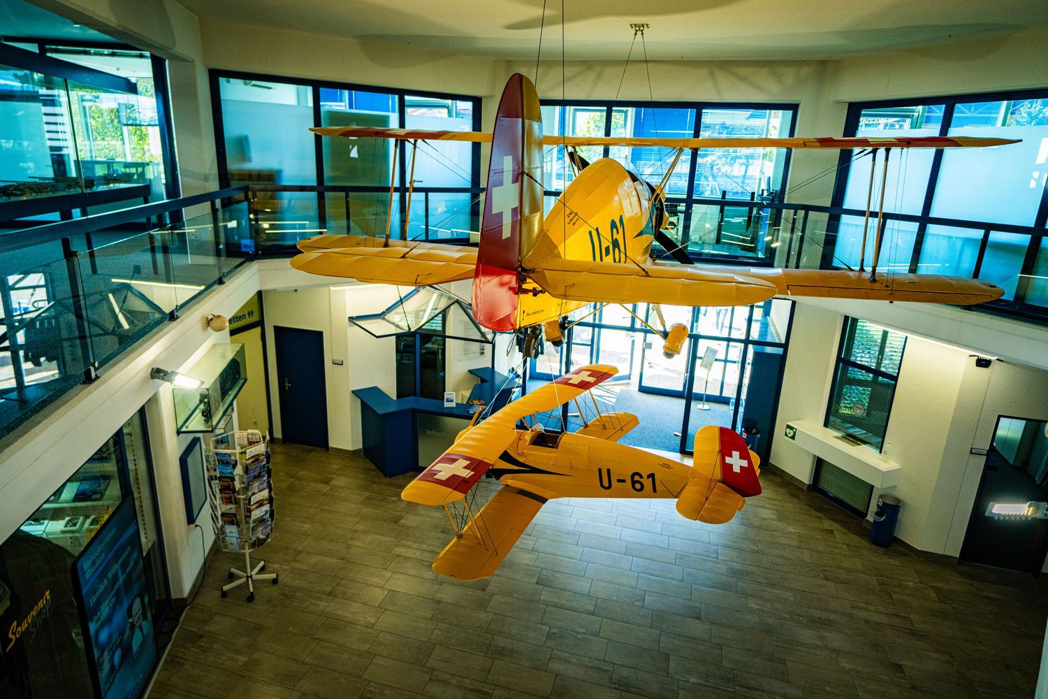 Two vintage yellow Swiss biplanes suspended from the ceiling of a bright museum hall with large windows and a wooden floor.