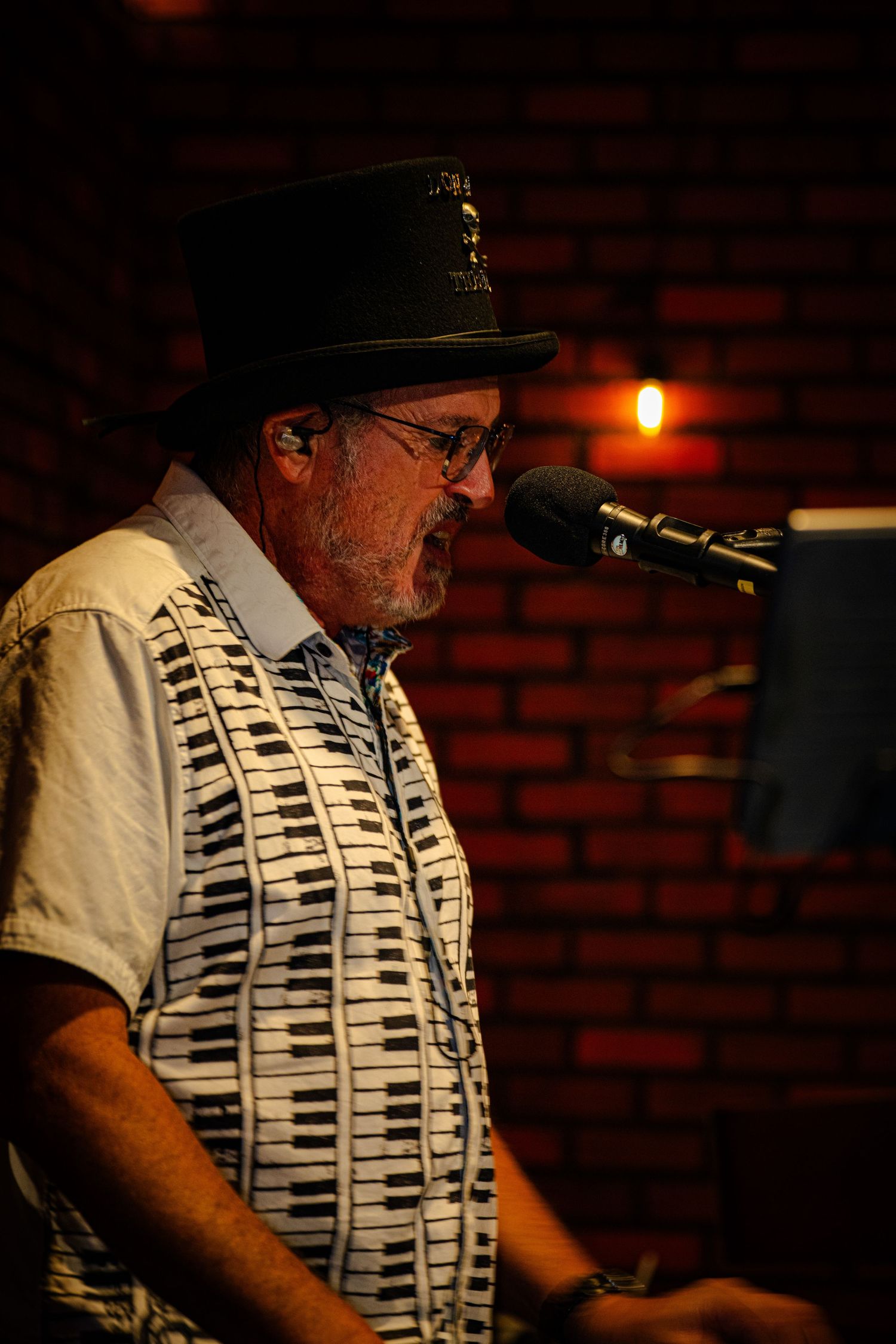 Musician in piano key patterned shirt and hat performs at a dimly lit brick-walled venue.