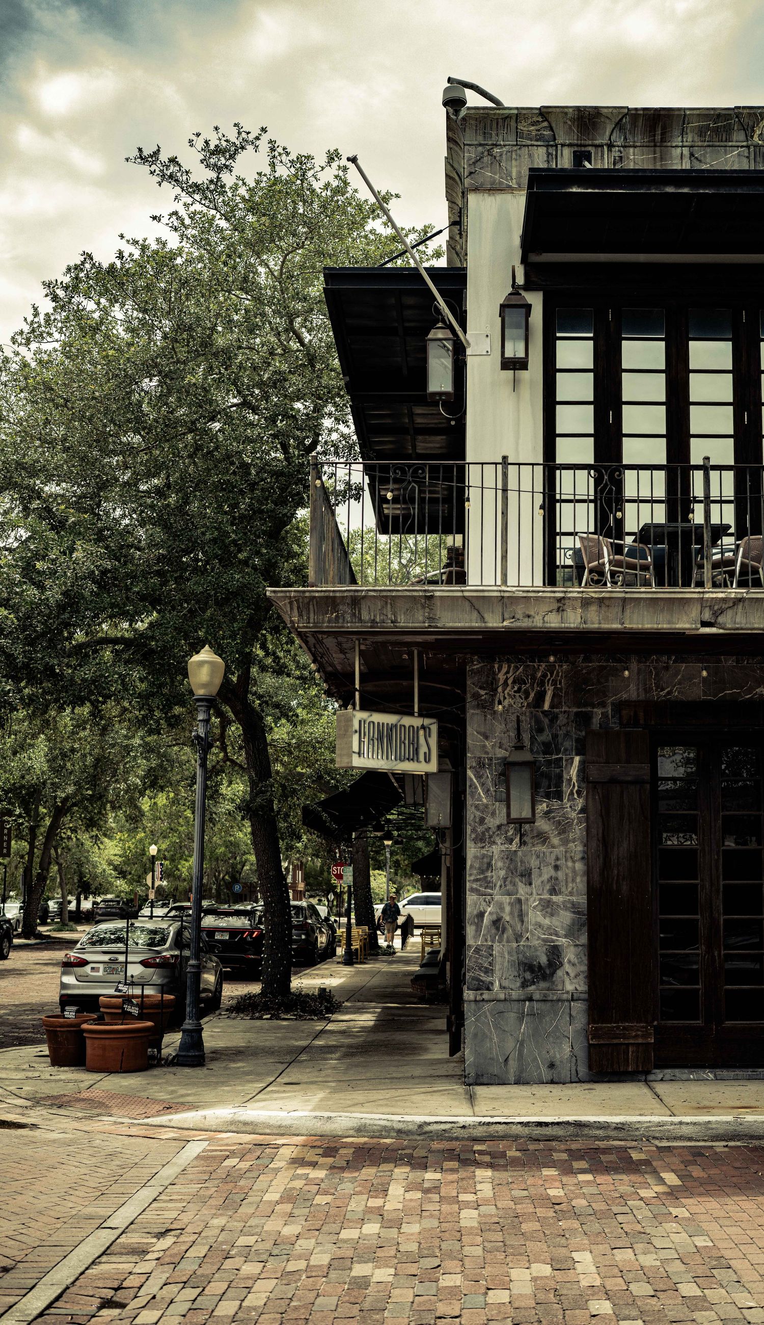 Historic corner building with wrought iron balcony, brick sidewalk, street lamp, and lush trees in a charming Southern downtown district.