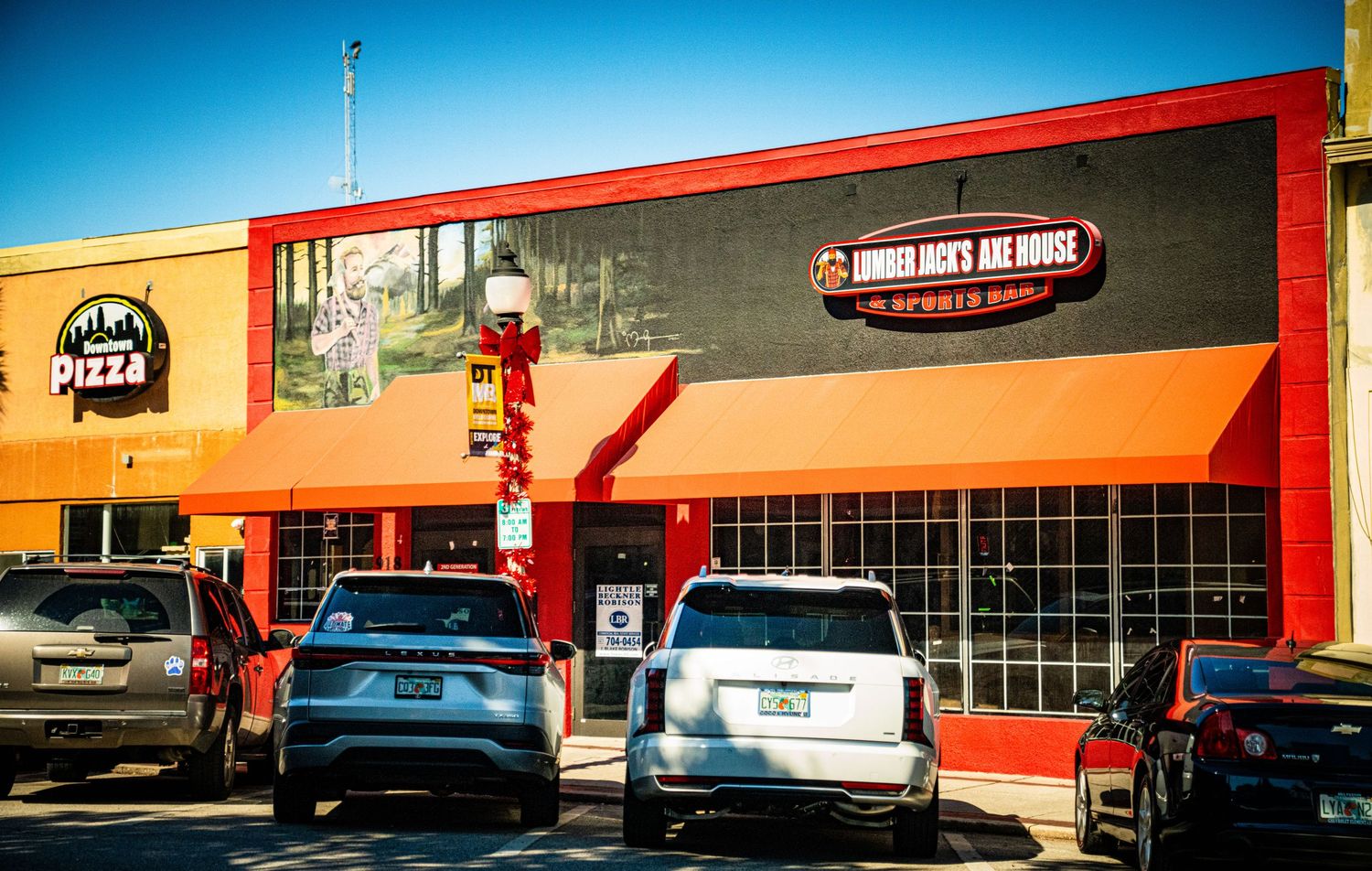 A long panoramic view of a strip mall exterior with orange and red trim and parked cars in front during daytime.