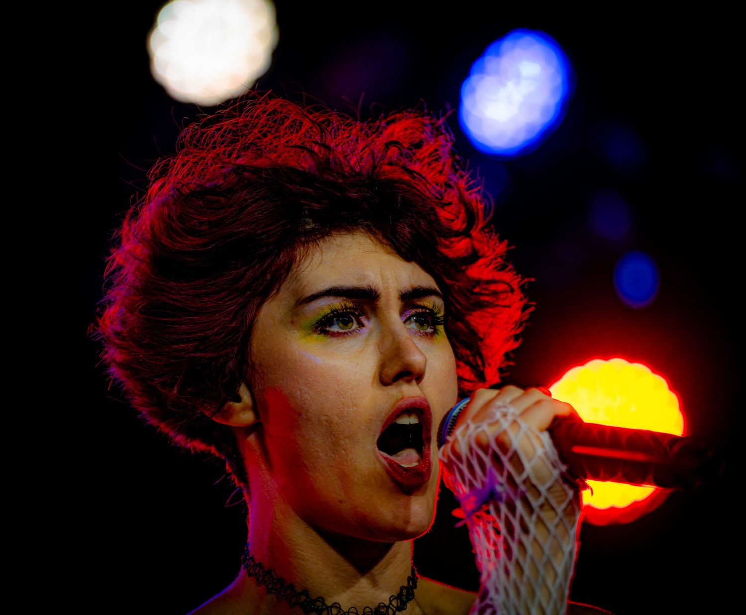 A performer singing passionately into a microphone under dramatic red and blue stage lights.