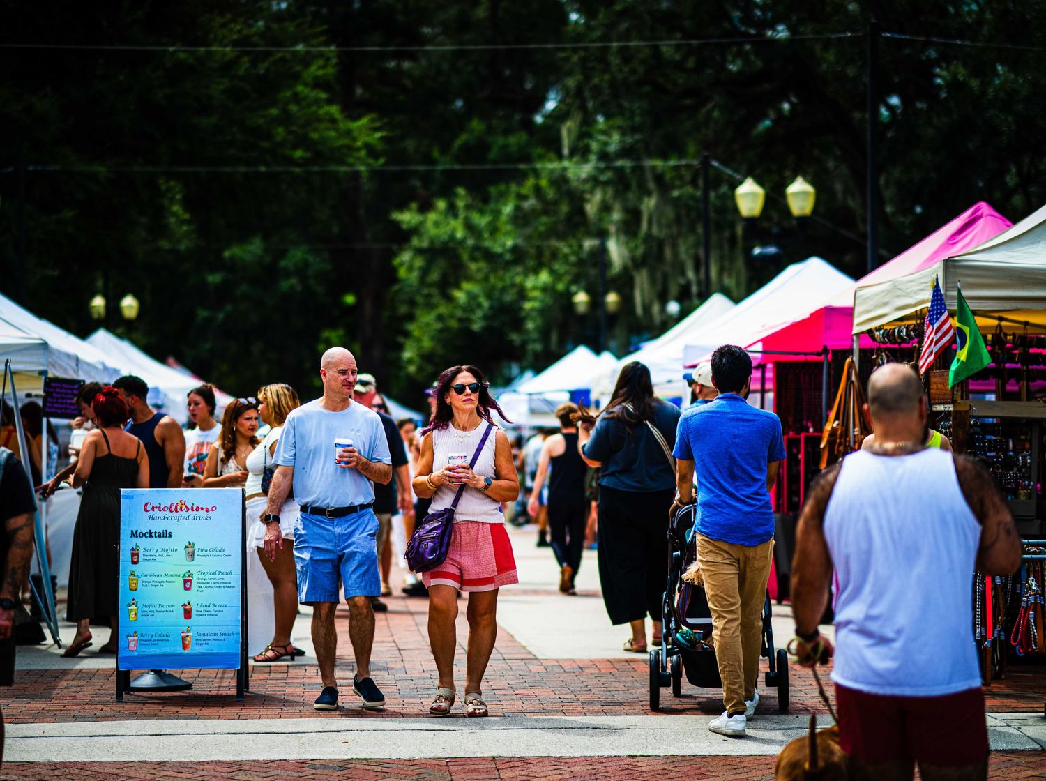 Crowds of people walking through an outdoor market with white tents on a sunny day.