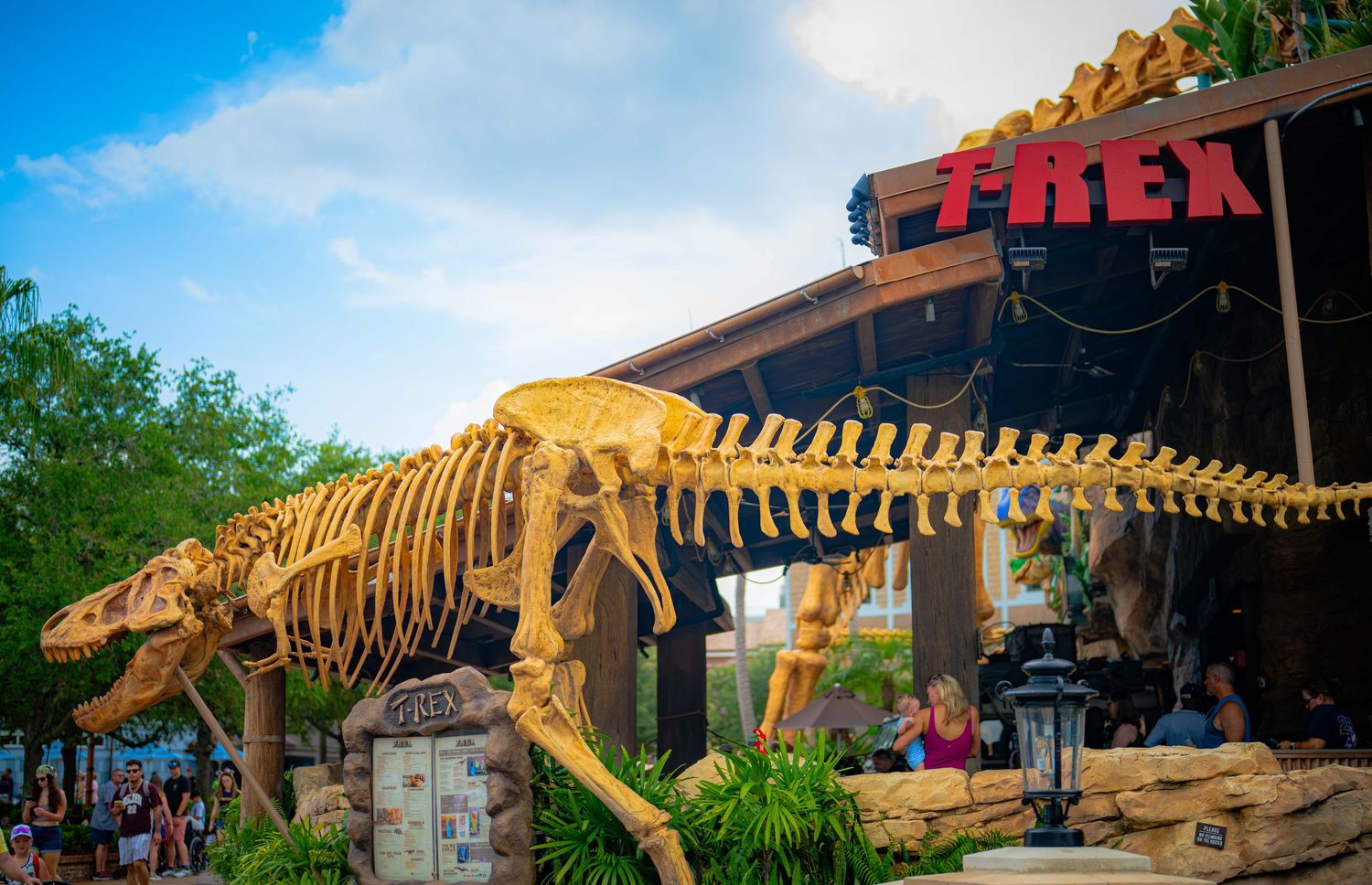 T-Rex skeleton replica displayed outside the T-Rex restaurant at Disney Springs, Florida, under a blue sky.