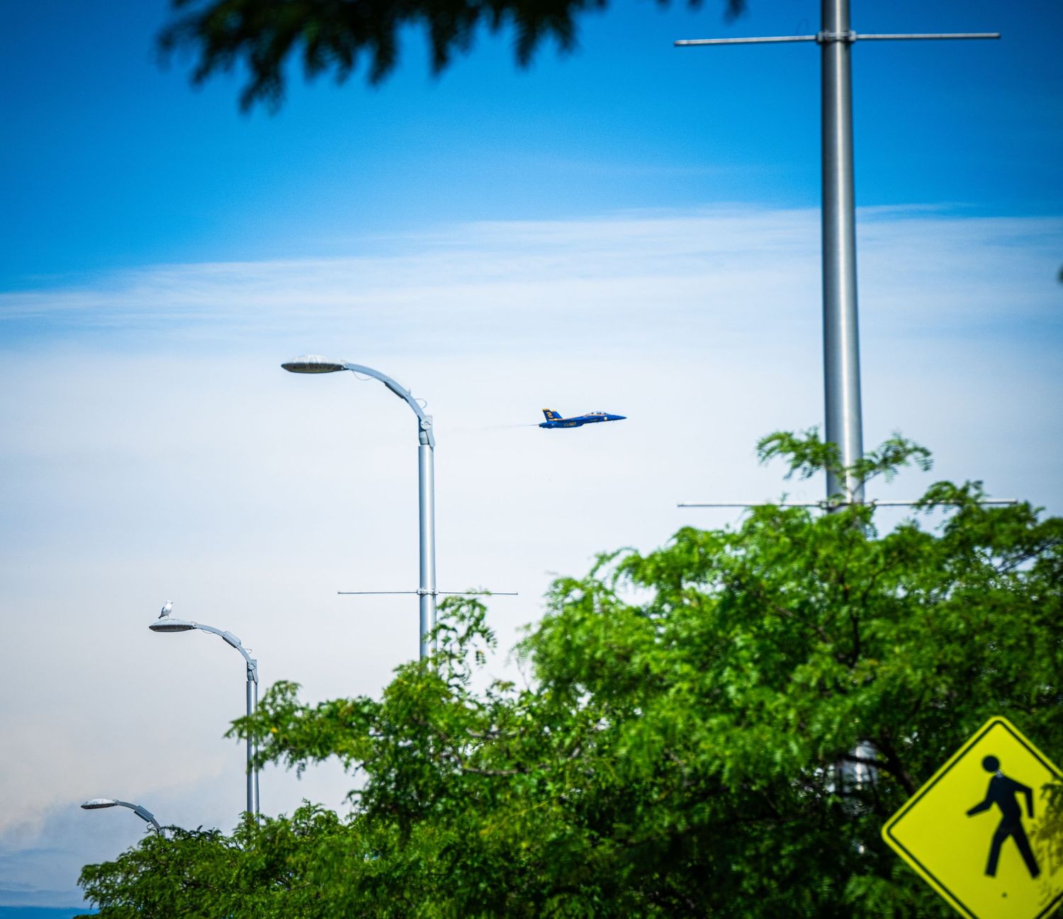 Military aircraft flying over street with pedestrian crossing sign and lamp posts.