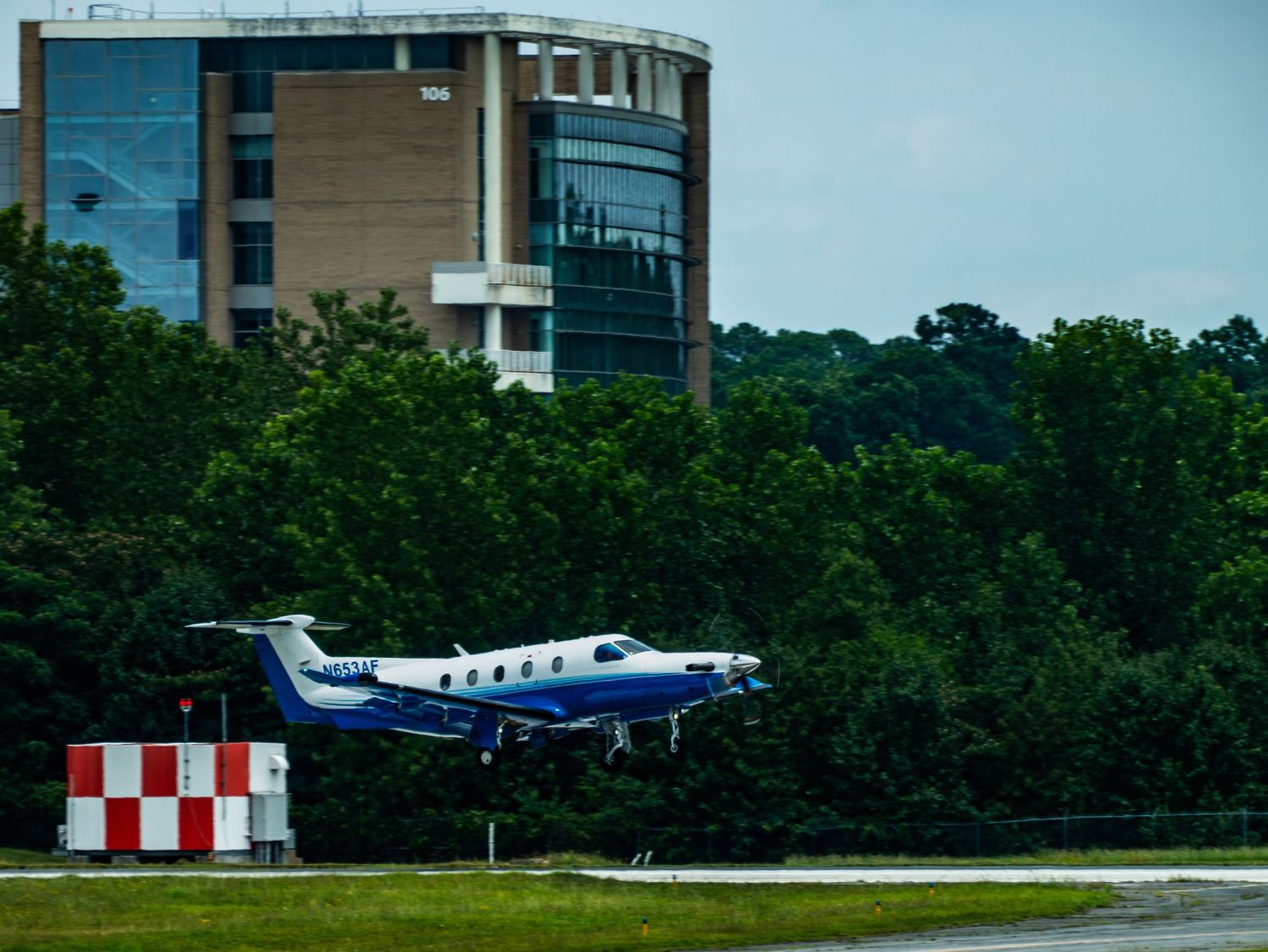 Small blue and white turboprop plane landing near a building surrounded by lush green trees.