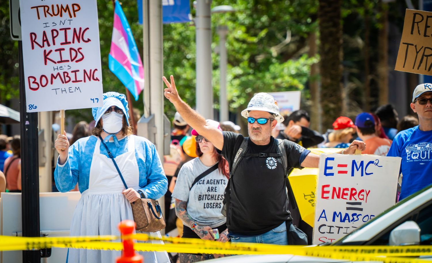 Protesters hold signs and flags at a political demonstration with yellow caution tape visible in the foreground.