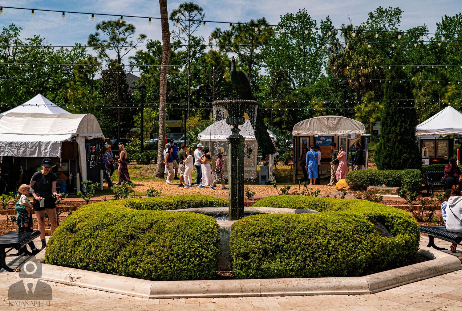 White vendor tents lined up at an outdoor art festival with trees in the background.